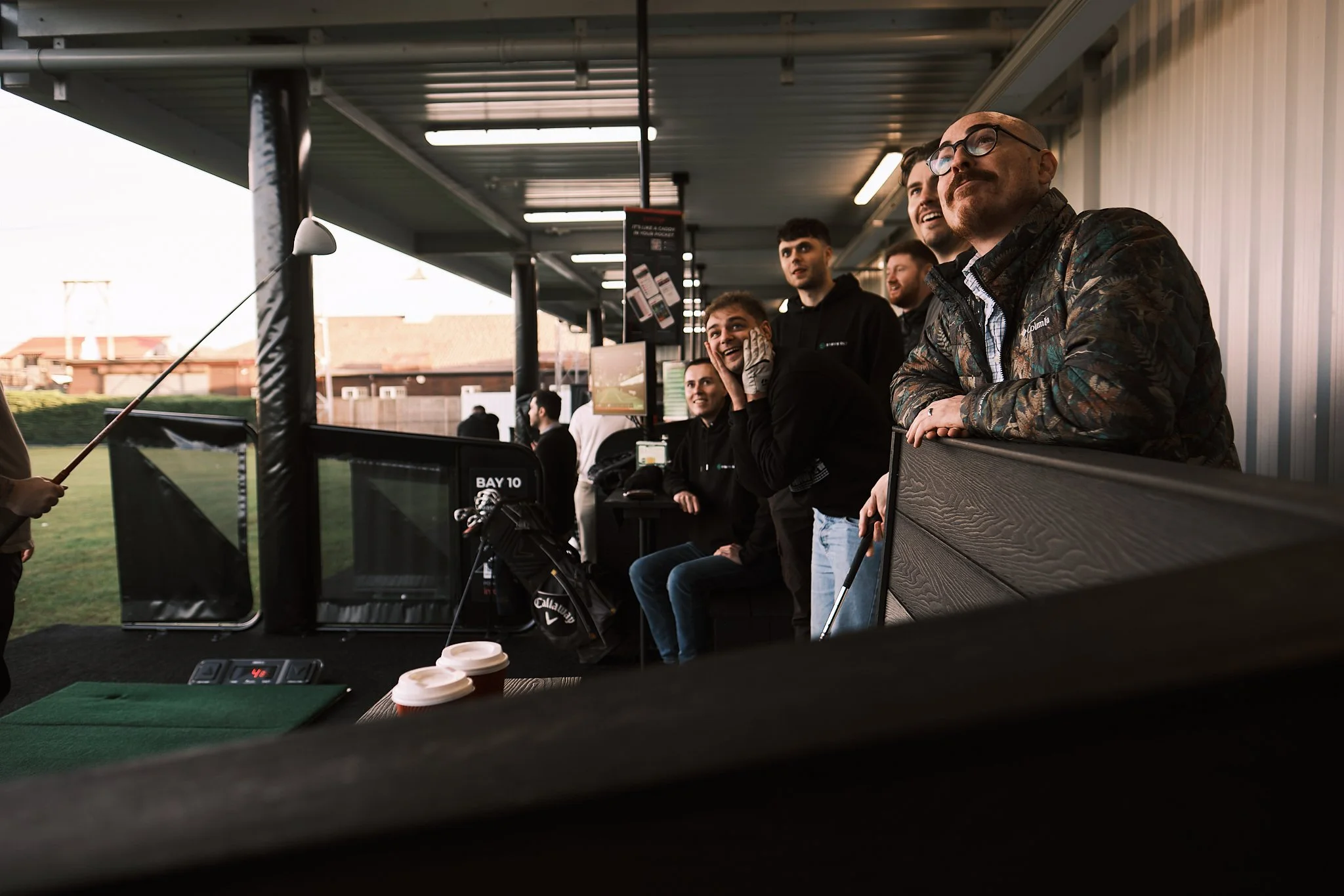 A group of people watching a golfer tee off at a golf driving range, some smiling and looking engaged.