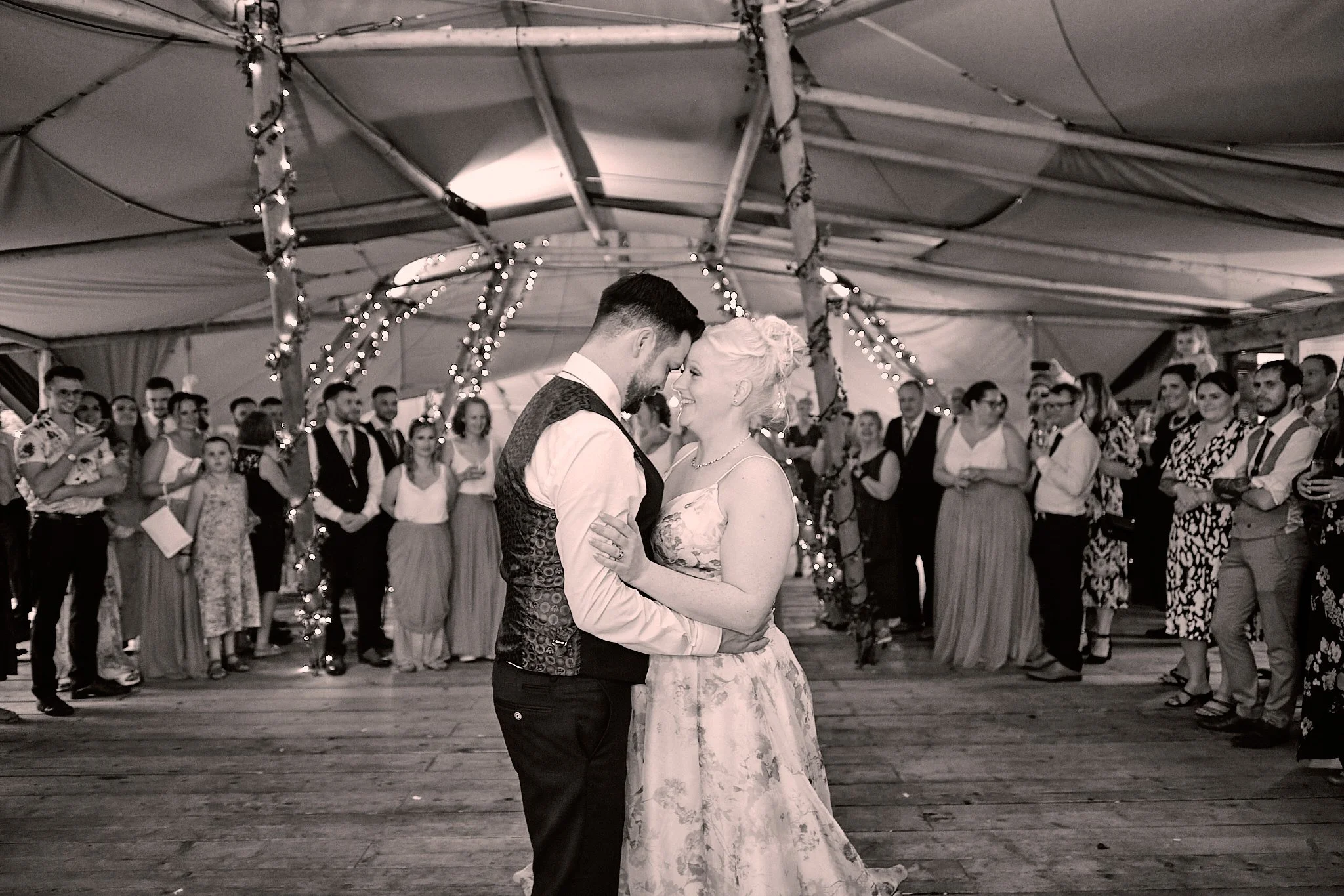 A couple dancing closely at a wedding reception, surrounded by guests under a decorated tent with string lights, in black and white.