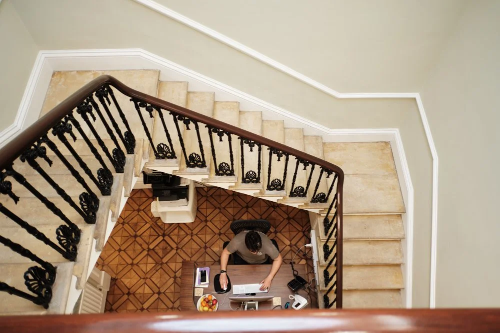 Overhead view of a person working at a desk on a staircase landing with a black ornate railing, a computer, and a bowl of fruit.