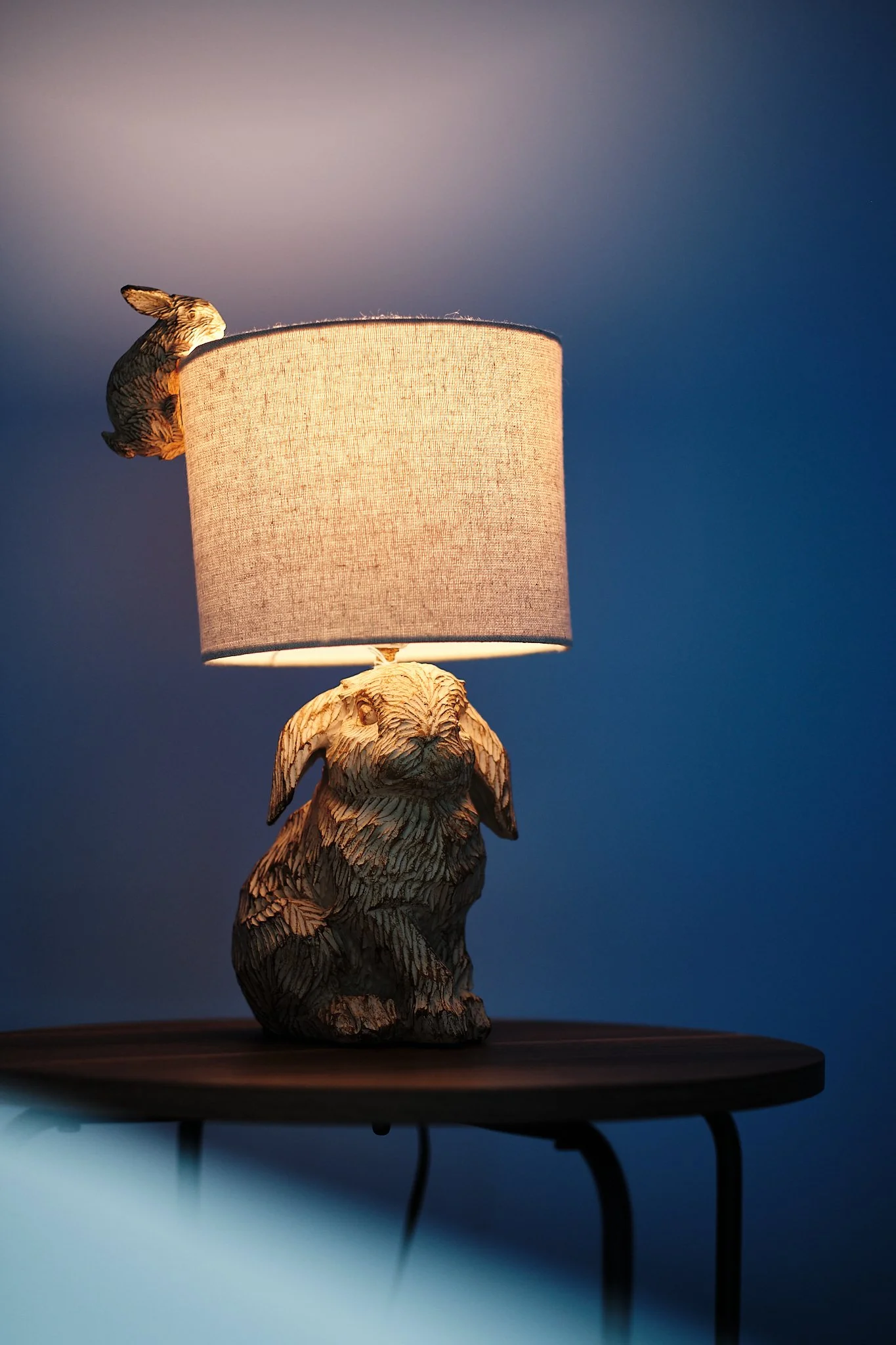 A table lamp with a base shaped like a sitting dog, with a lampshade on top, and a decorative bird attached on the side of the lampshade, placed on a dark wooden table against a dark background.