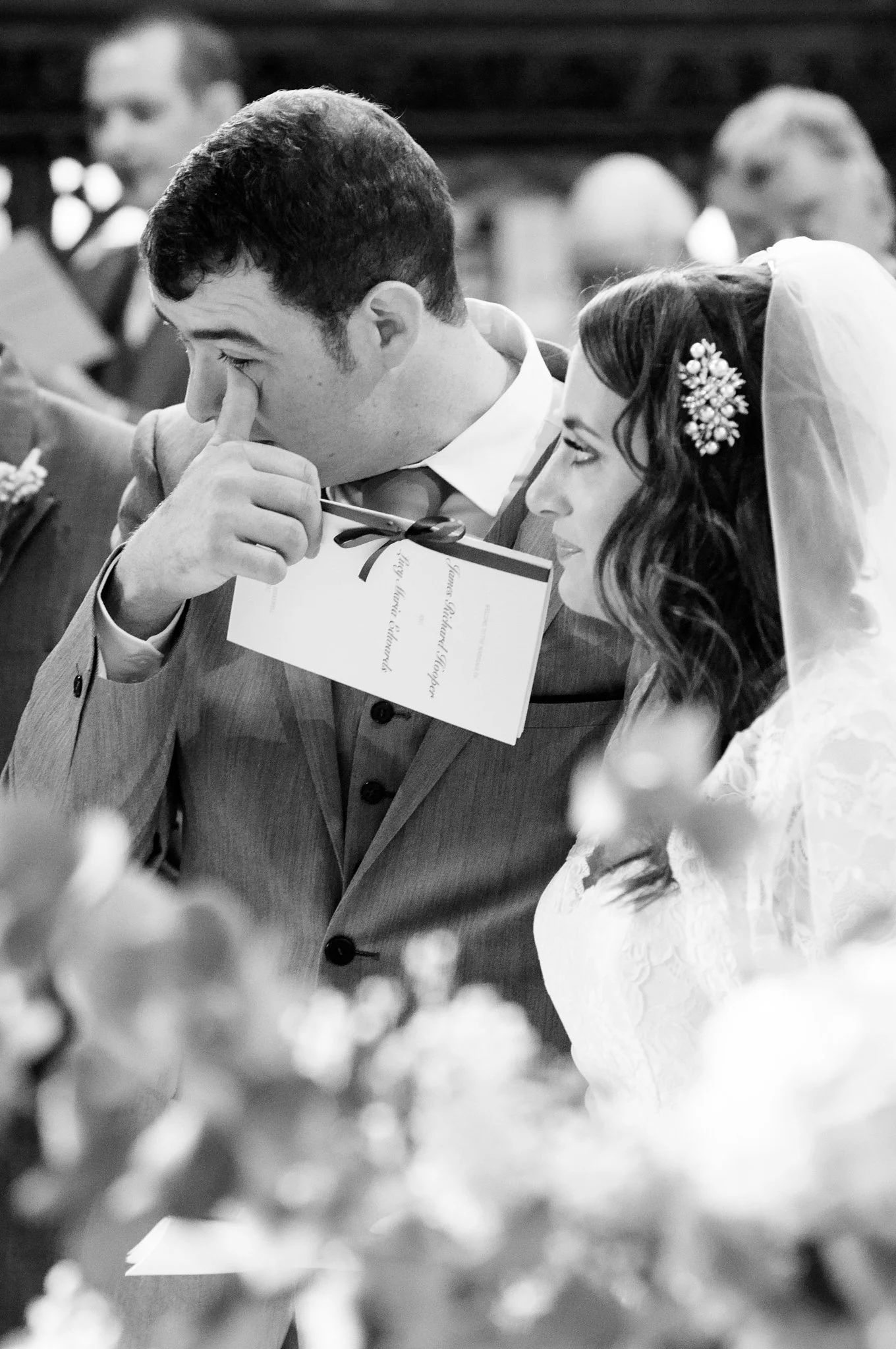 A black-and-white photo of a wedding ceremony showing a man and woman, possibly the bride and groom, in close interaction. The man has dark hair and is wearing a suit, holding a small object near his face, and the woman has dark hair with a decorativ