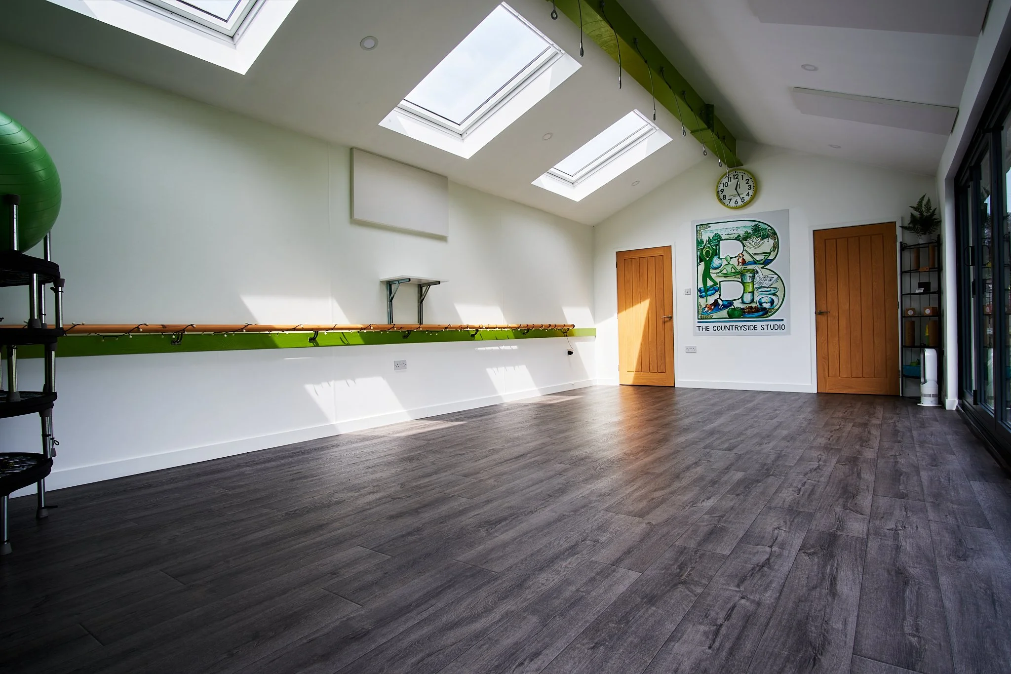 Empty fitness or dance studio with dark wood flooring, white walls, three skylights, two wooden doors, a painting, clock, black shelving unit, and some green gym equipment.