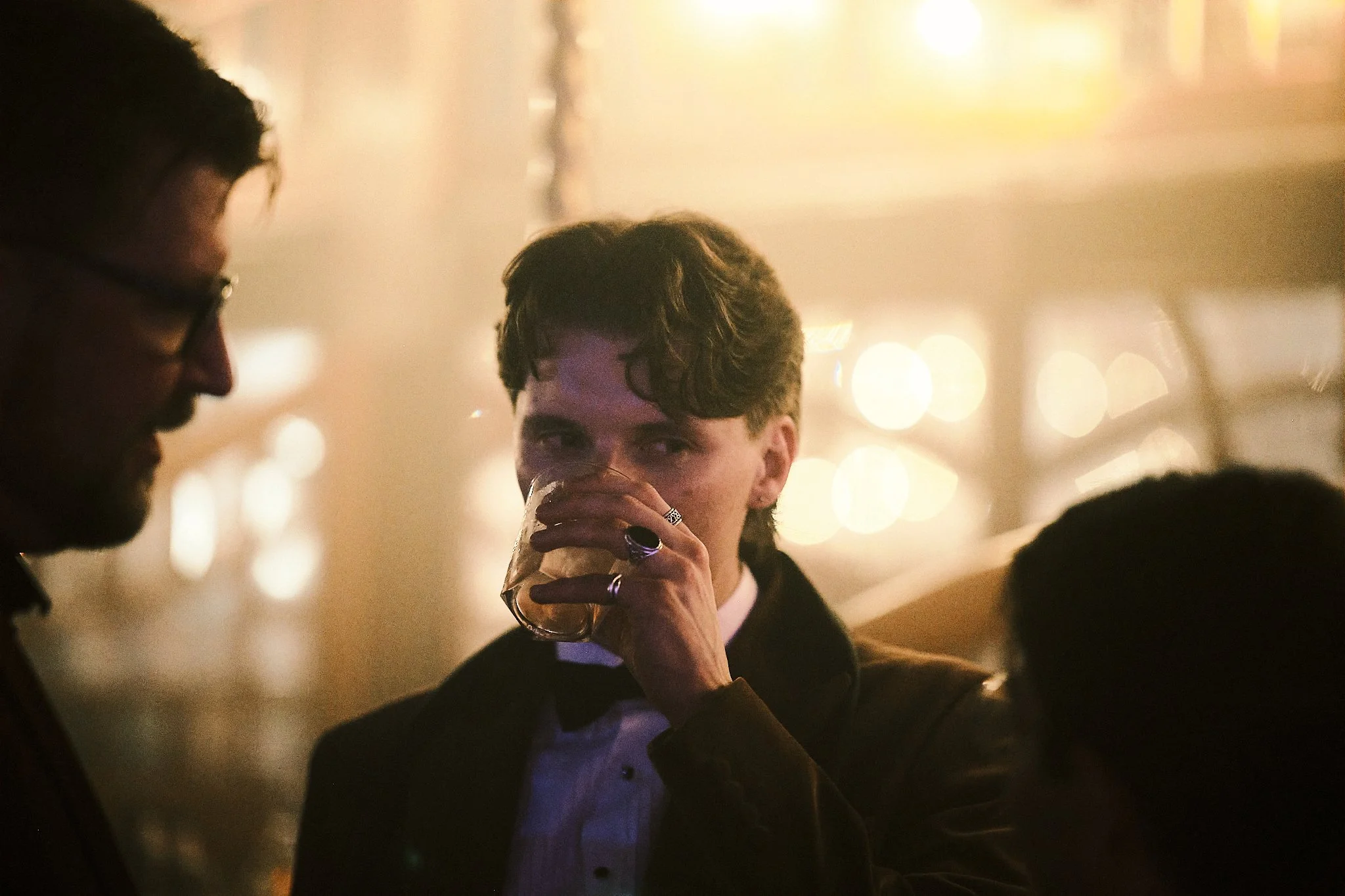 Three men in formal attire socializing at a dimly lit event, with the man in the center drinking from a glass and the other two standing nearby.