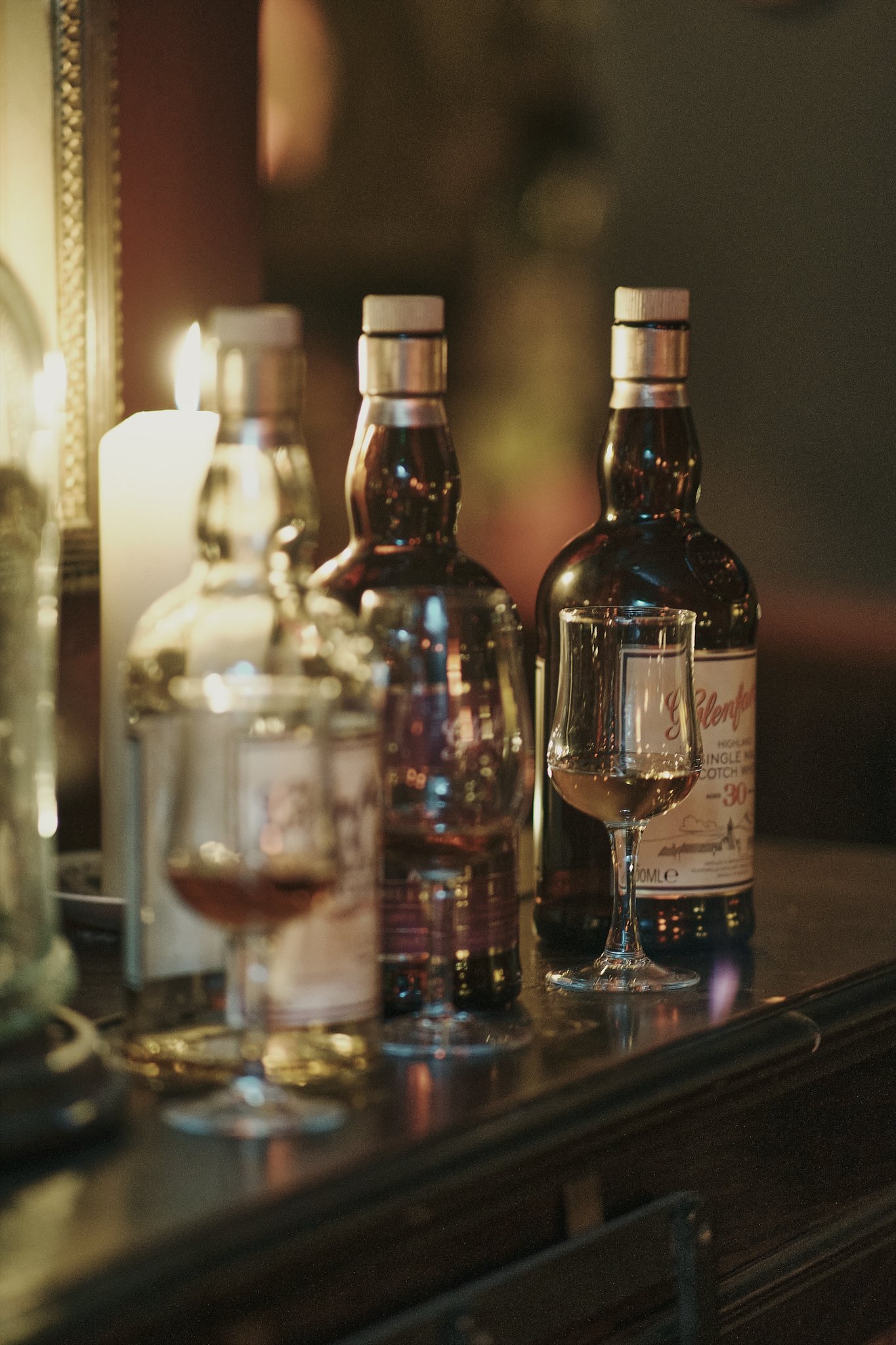 Multiple bottles of Scotch whisky and glasses of whisky on a dark wooden bar in a cozy, dimly lit setting with a candle in the background.
