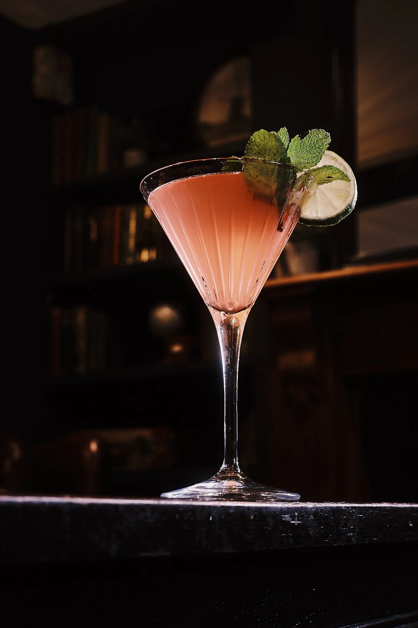 A pink cocktail in a martini glass garnished with a slice of lime and fresh mint leaves.