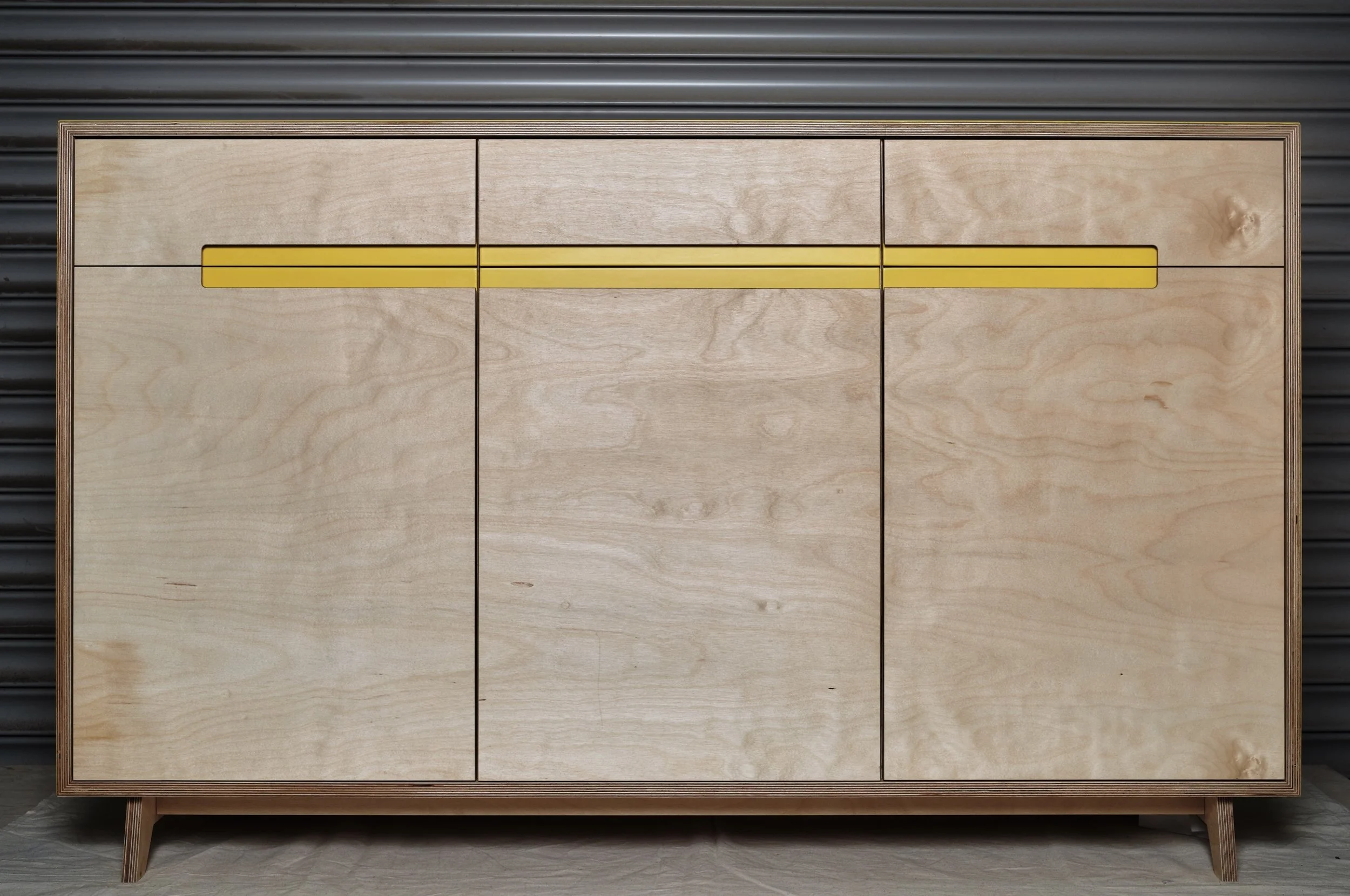 A minimalist wooden cabinet with a light finish, featuring three yellow horizontal lines on the top section, set against a dark industrial wall background.