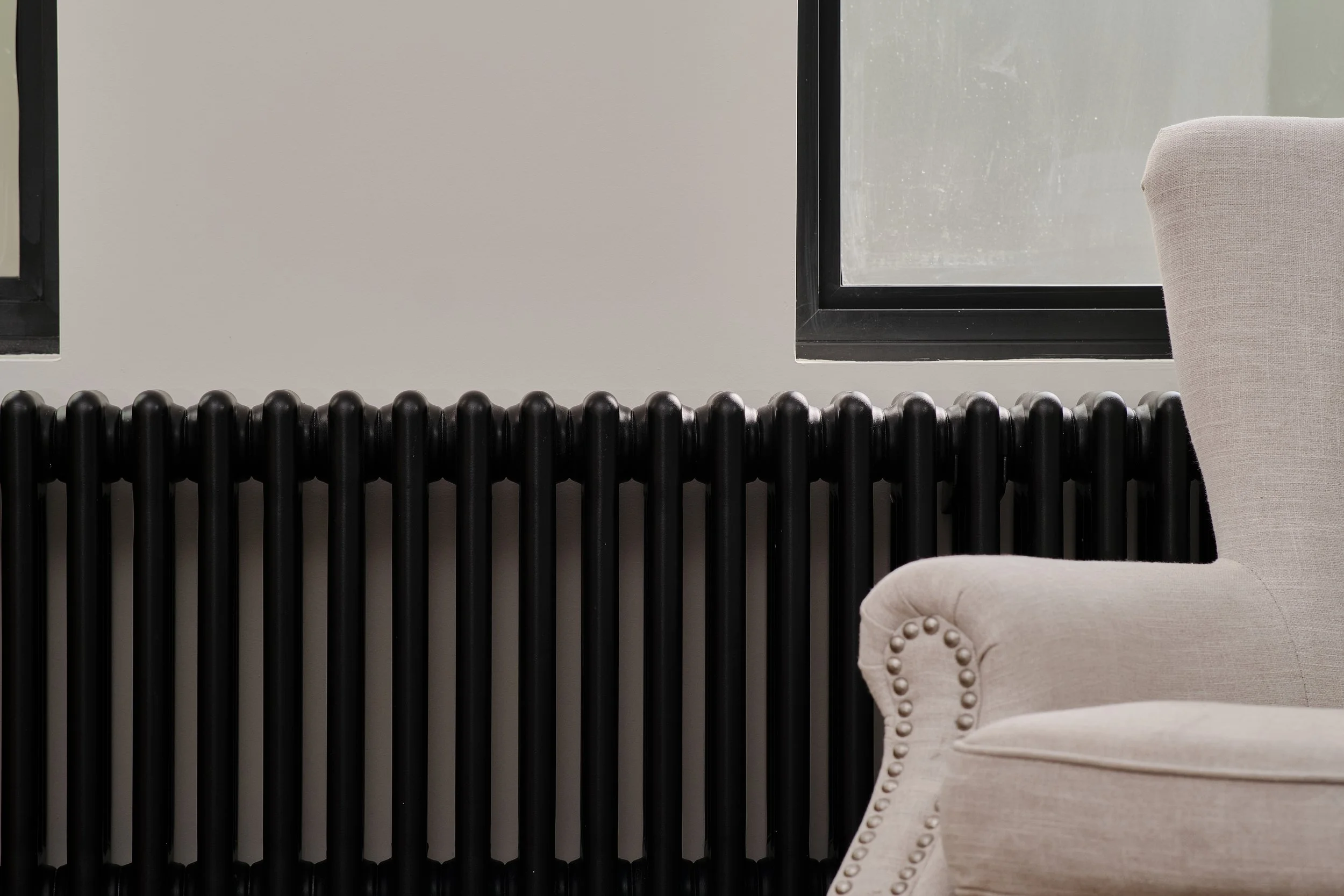 Part of a beige upholstered armchair with nailhead trim, a black radiator, and a window with a black frame, in a minimalistic interior setting.