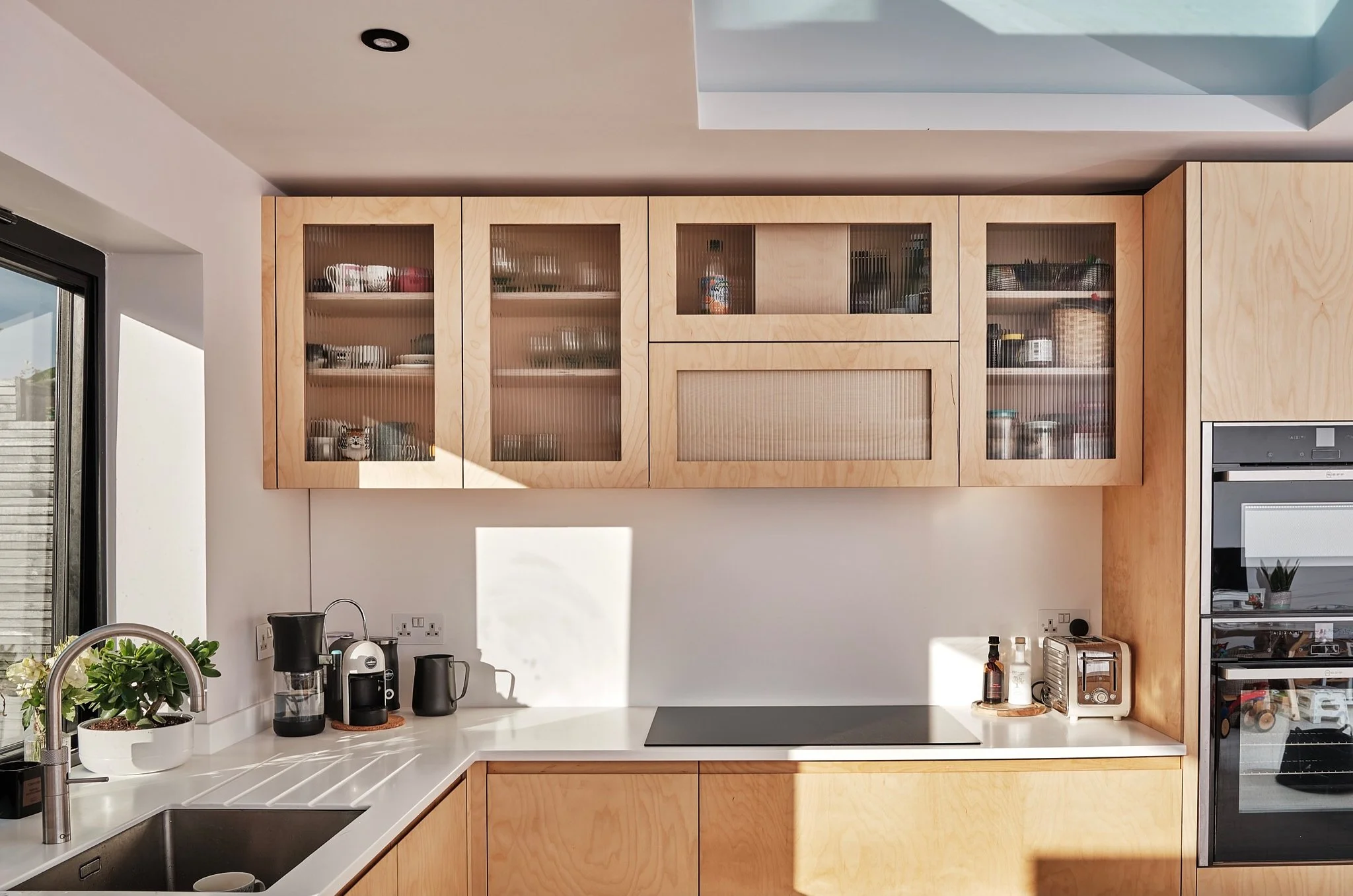 Modern kitchen with natural wood cabinets, white countertops, a black stovetop, and small appliances, illuminated by natural sunlight.
