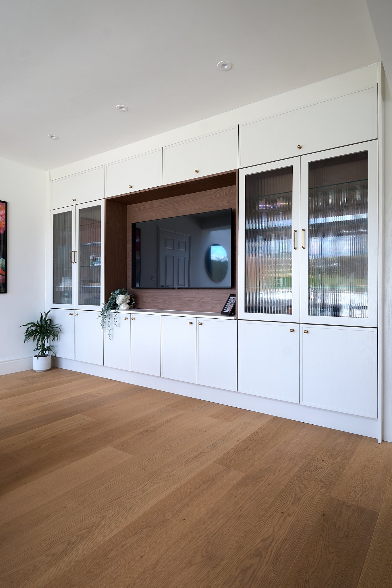 Living room entertainment center with a wall-mounted flat-screen TV, white cabinets with gold knobs, glass-paneled doors, a potted plant, and decorative items.