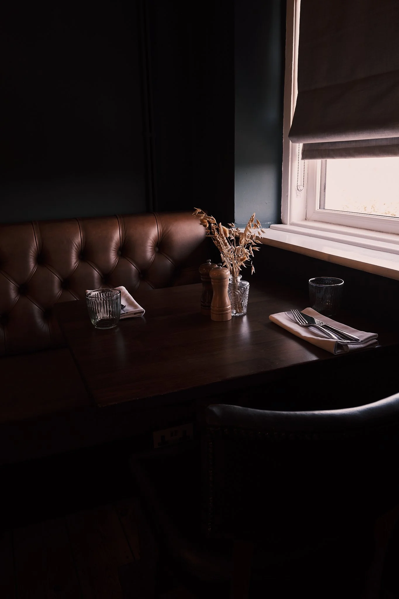 A cozy restaurant table near a window with a brown leather tufted bench, a dark wooden table set with a napkin, utensils, glasses, and a small vase with dried flowers, illuminated by natural light.