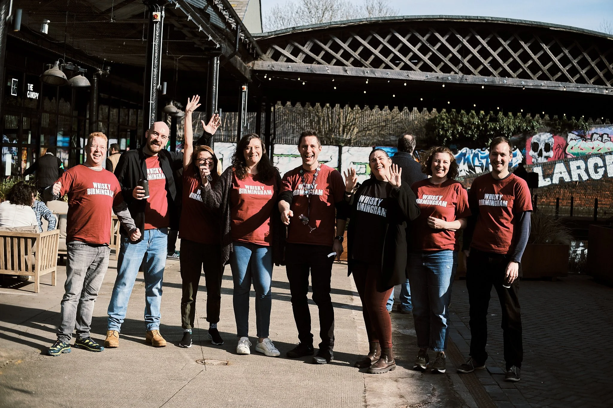 Group of nine people standing outside, smiling and waving, wearing matching red shirts that say "Whisky Birmingham" on a sunny day.