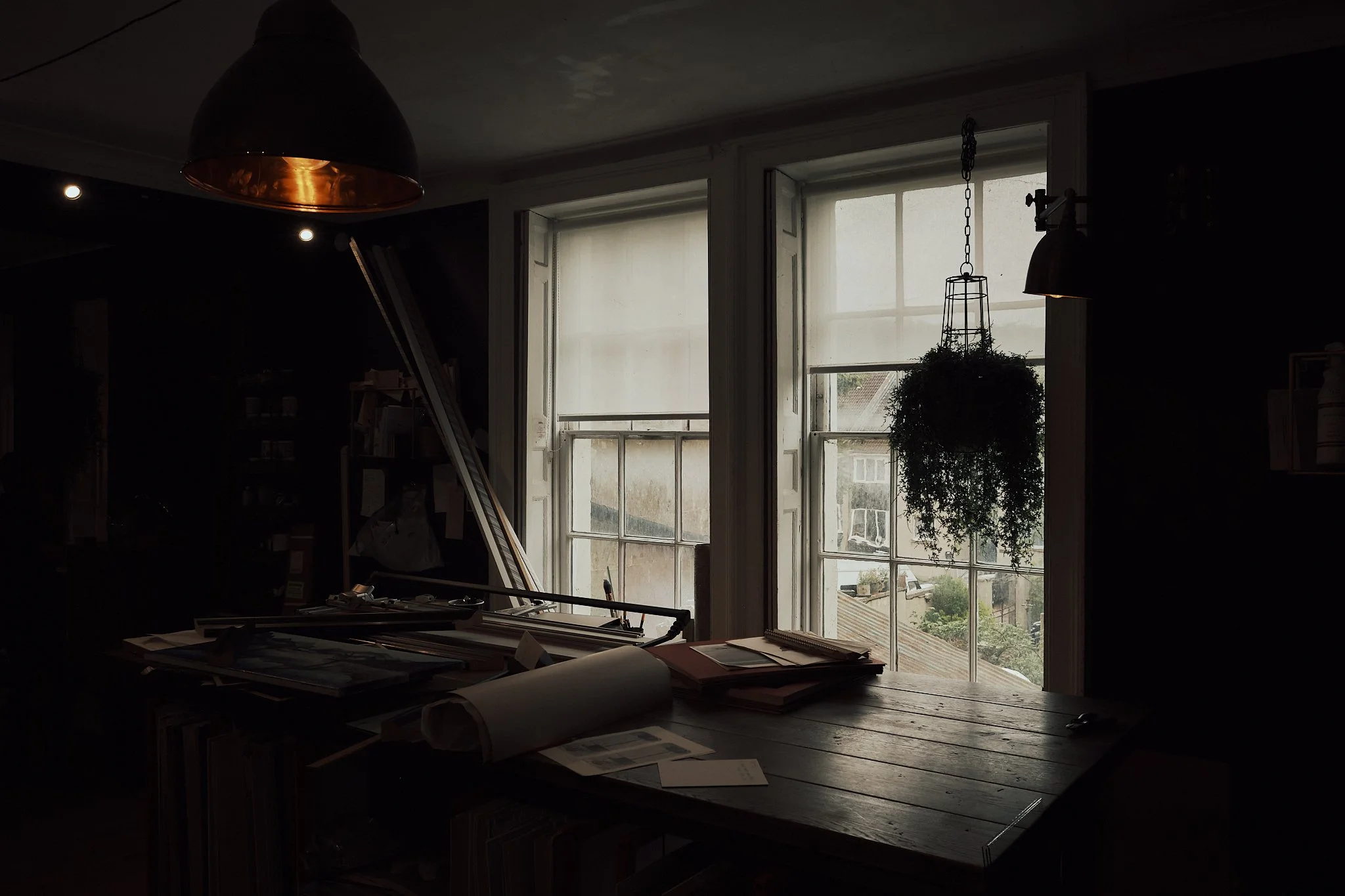 A dimly lit room with two large windows, a hanging plant, and a wooden table with papers and rolled-up documents.