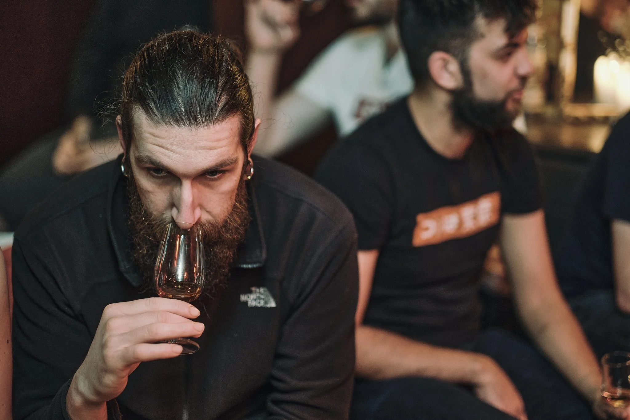 A bearded man with long hair in a ponytail holding a glass of wine to his nose, sitting among other people in a dimly lit setting.