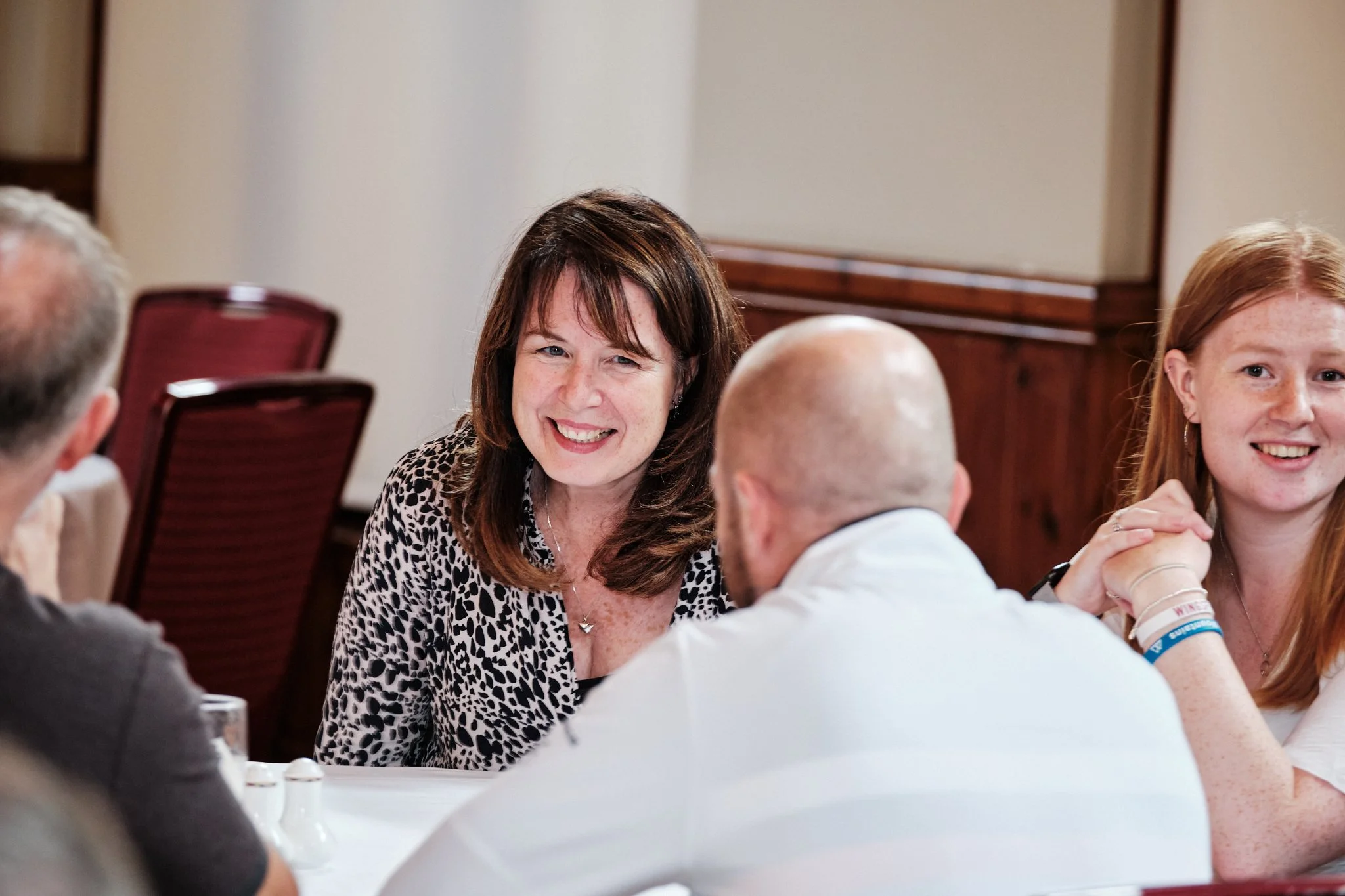 A group of people sitting at a round table engaged in conversation, including a woman smiling at the camera, a young woman with red hair, and two men with blurred faces.