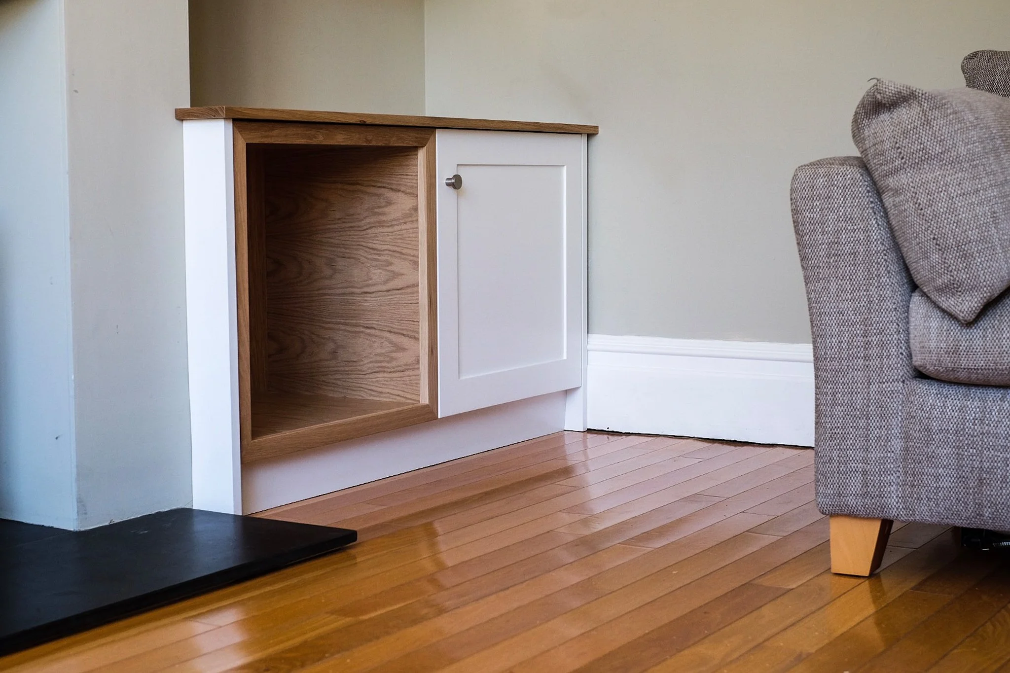Corner of a room with hardwood floor, partial view of a gray upholstered sofa on the right, an empty wooden and white cabinet against the wall, and a black mat near the corner.