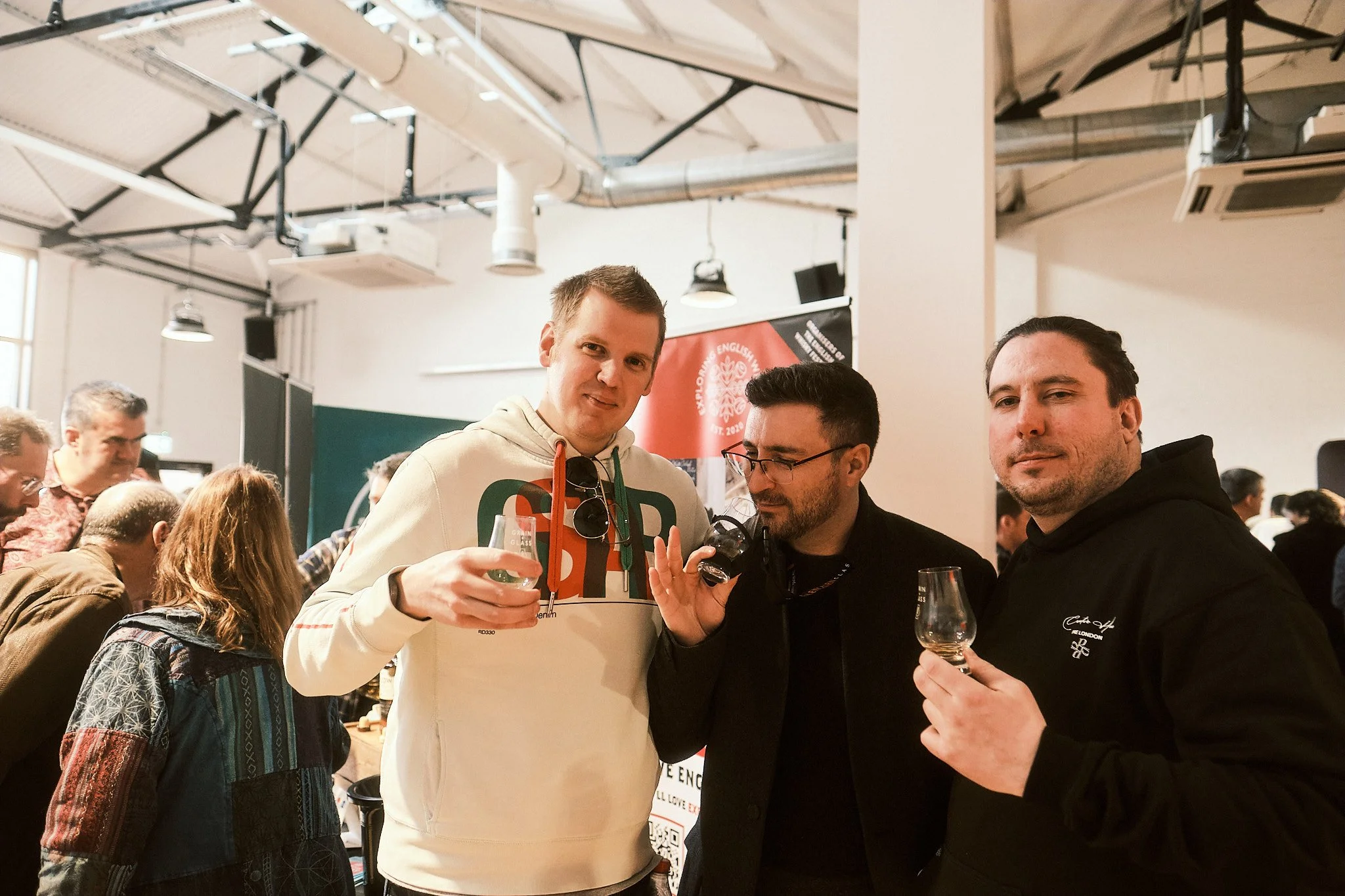 Three men holding glasses at an indoor alcohol tasting event, with a large group of people in the background.