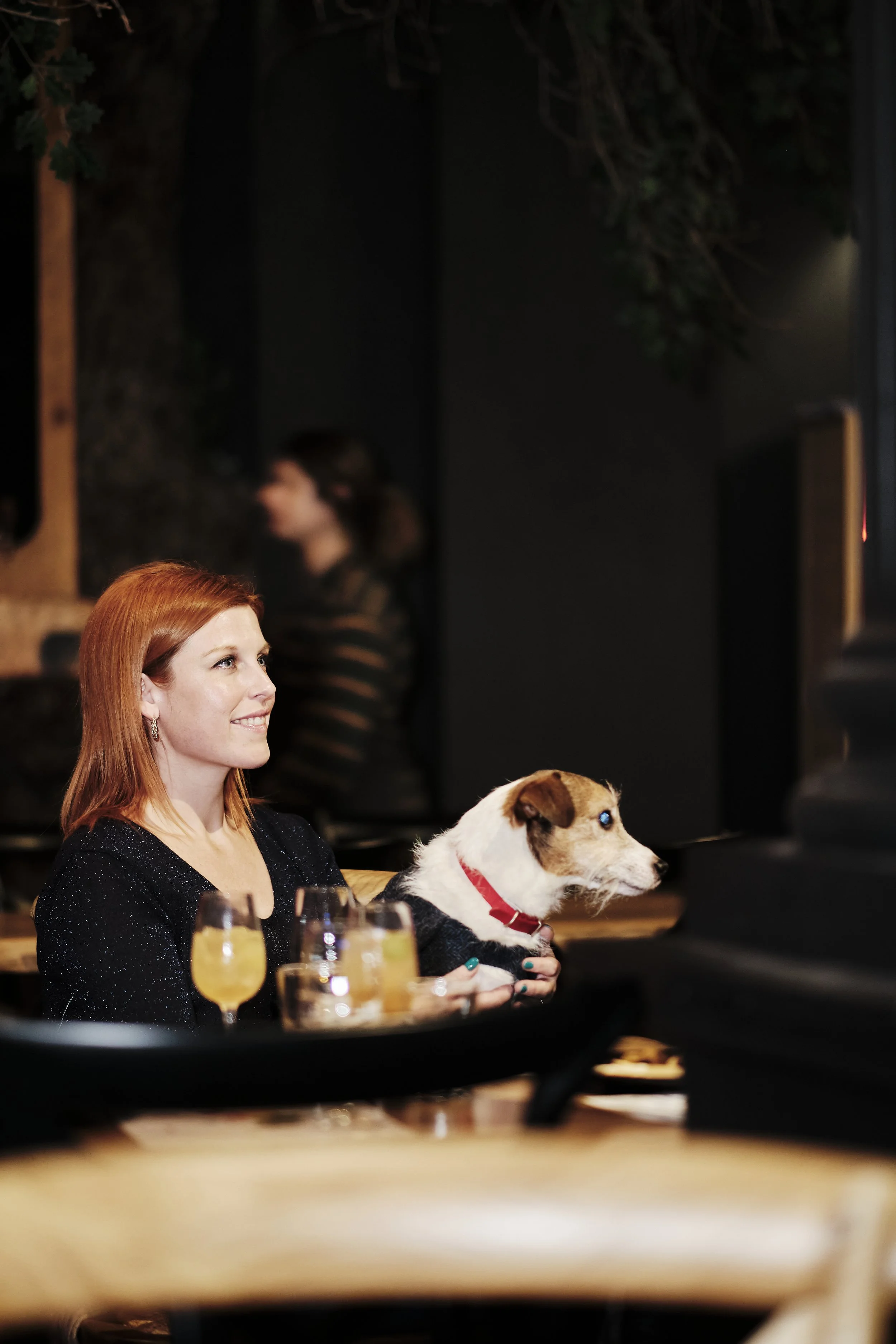 A woman with red hair smiling while holding a small dog, possibly a Jack Russell Terrier, with a red collar. They are sitting at a table with drinks and are watching a screen, with another person blurred in the background.