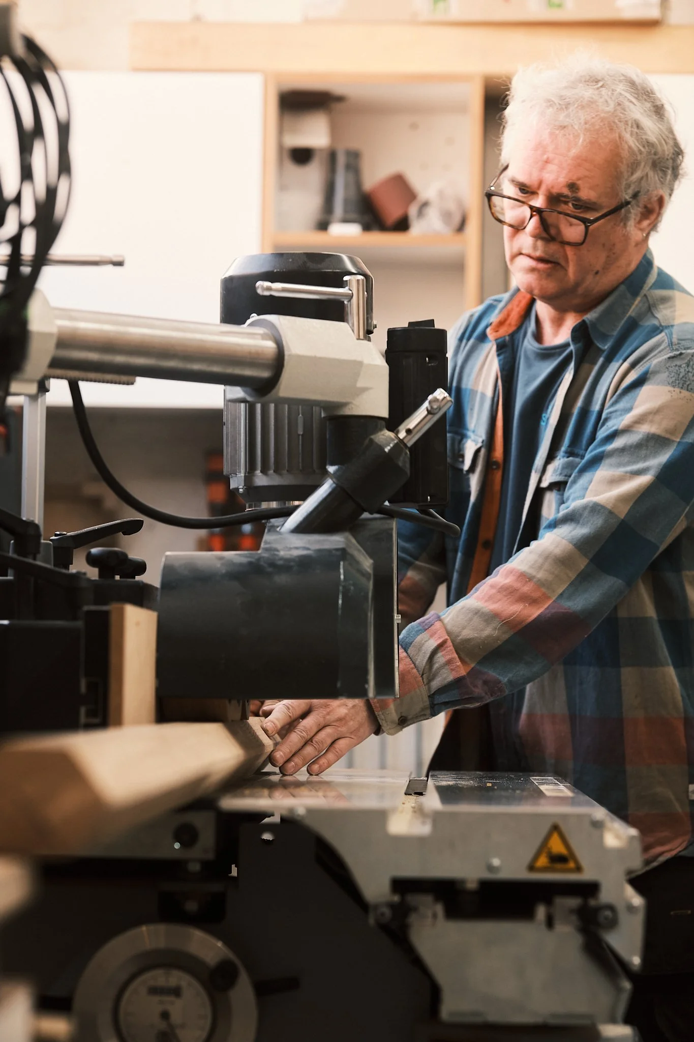 An older man with gray hair and glasses wearing a plaid shirt working with a machine in a workshop or woodworking shop.