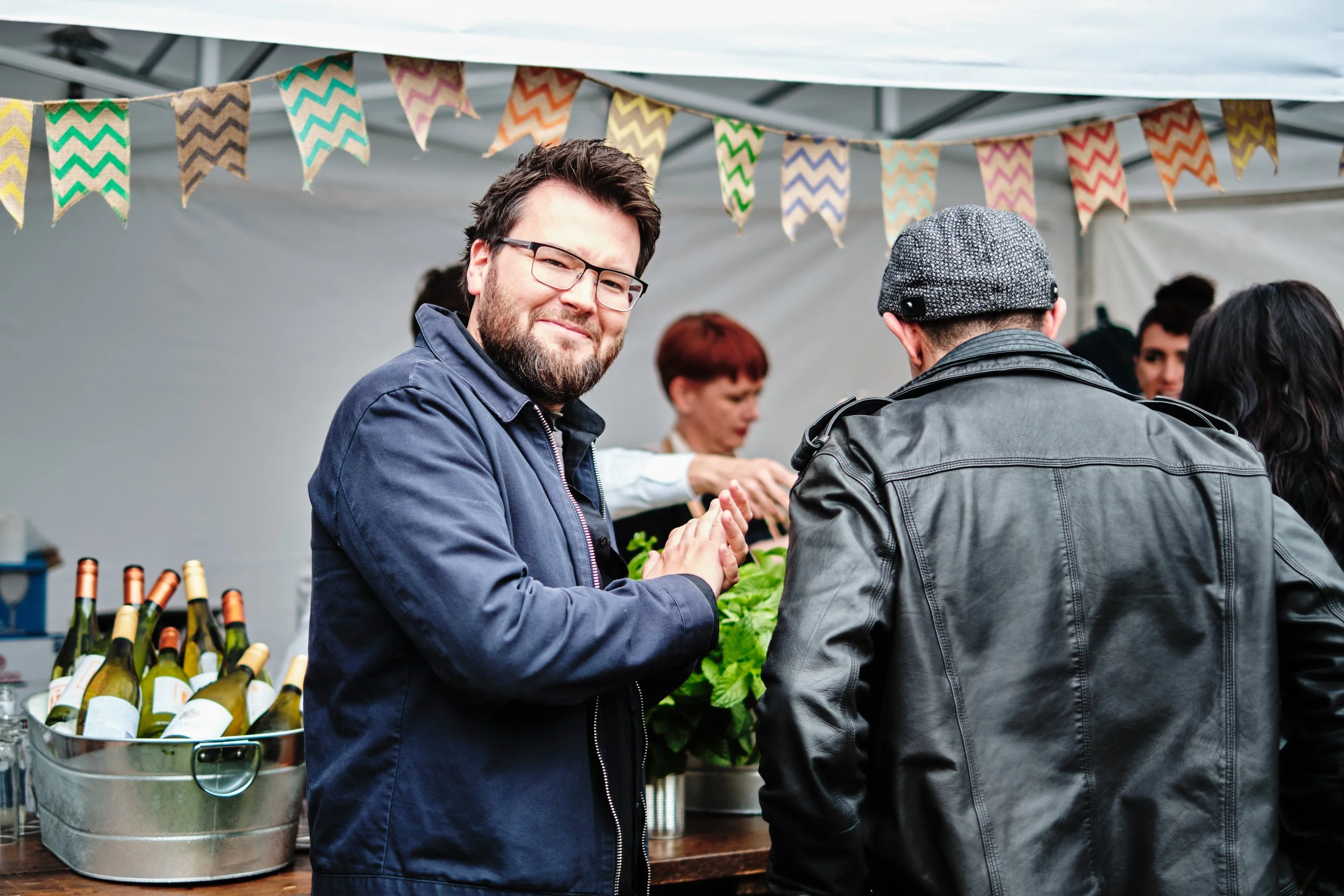 A man with glasses and a beard smiles at camera while standing at an outdoor event, with a crowd of people and bottles of wine on the table behind him.