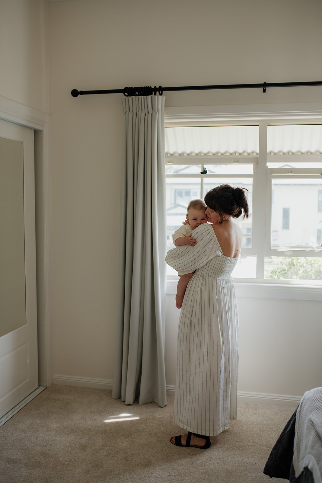 Gold Coast newborn photographer captures mother at home standing near window with baby during Gold Coast newborn photo shoot.