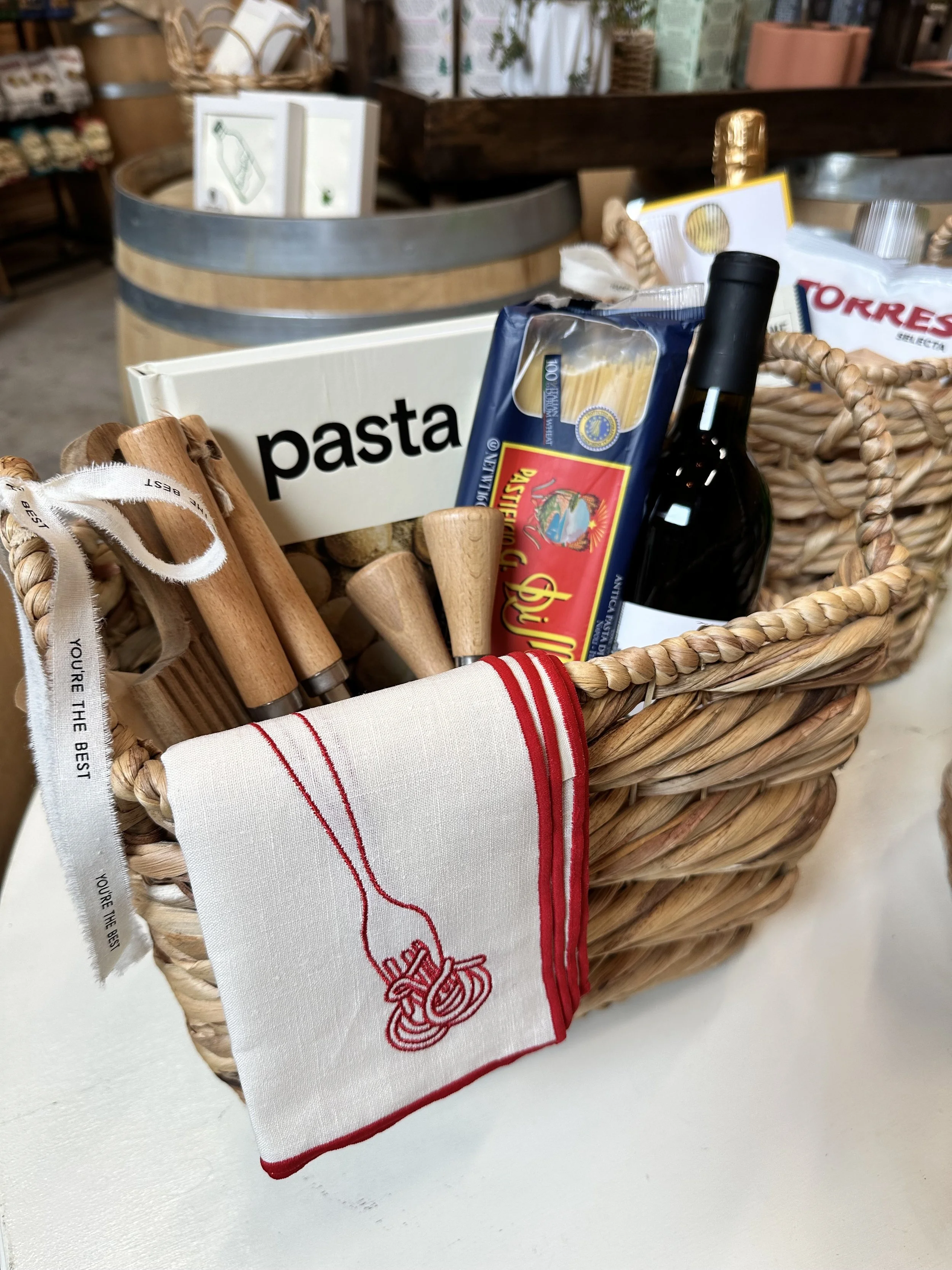 A wicker basket containing cooking utensils, a box of pasta, a bottle of wine, a package of spaghetti, and a red and white cloth with an embroidered fork design.