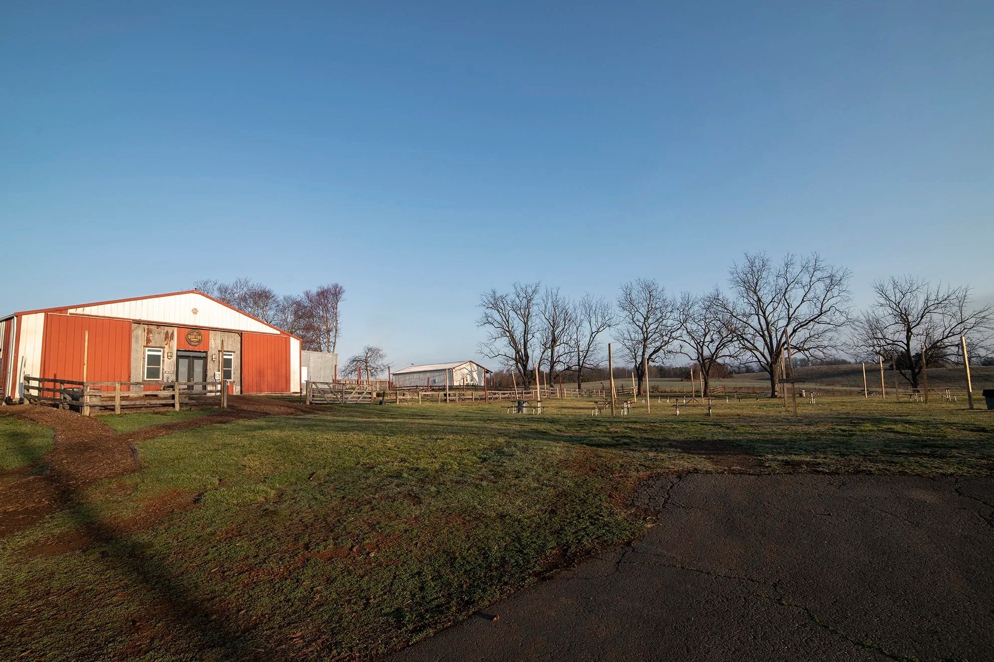 A rural farm scene with a large red barn, a white shed, leafless trees, grassy areas, fences, and a clear blue sky.