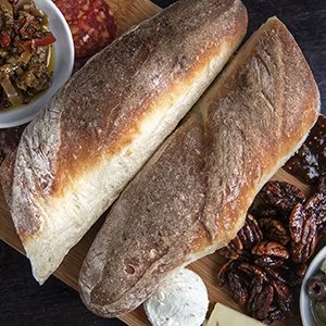 Two baguettes on a wooden cutting board with side dishes of pecans, a small bowl of jam, and a dollop of butter.