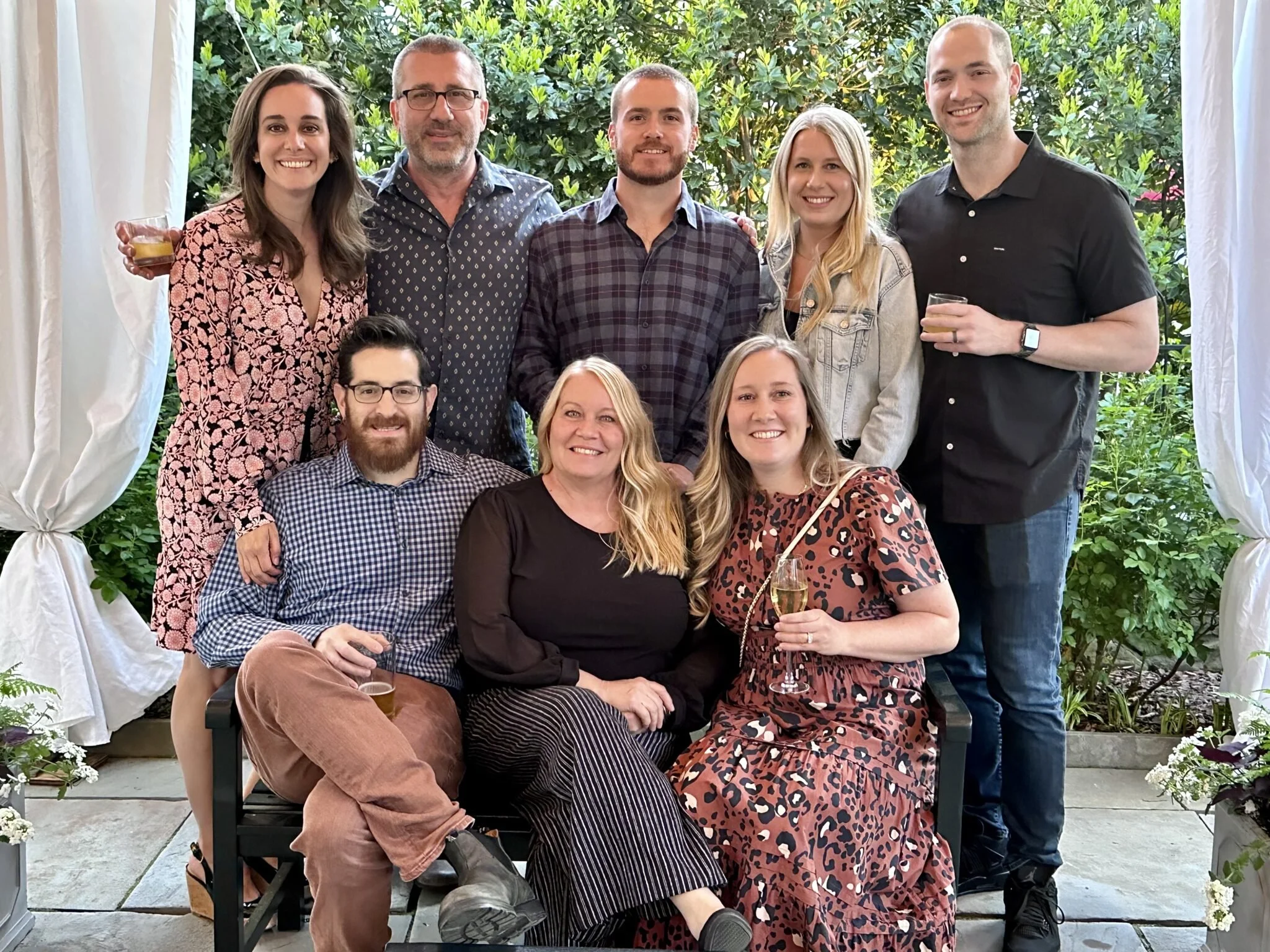 A group of nine people posing for a photo in a garden or backyard. They are smiling, dressed casually, some holding drinks, with greenery in the background and white curtains framing the scene.