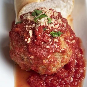 A meatball covered in marinara sauce, topped with grated cheese and garnished with fresh parsley, served on a piece of bread.