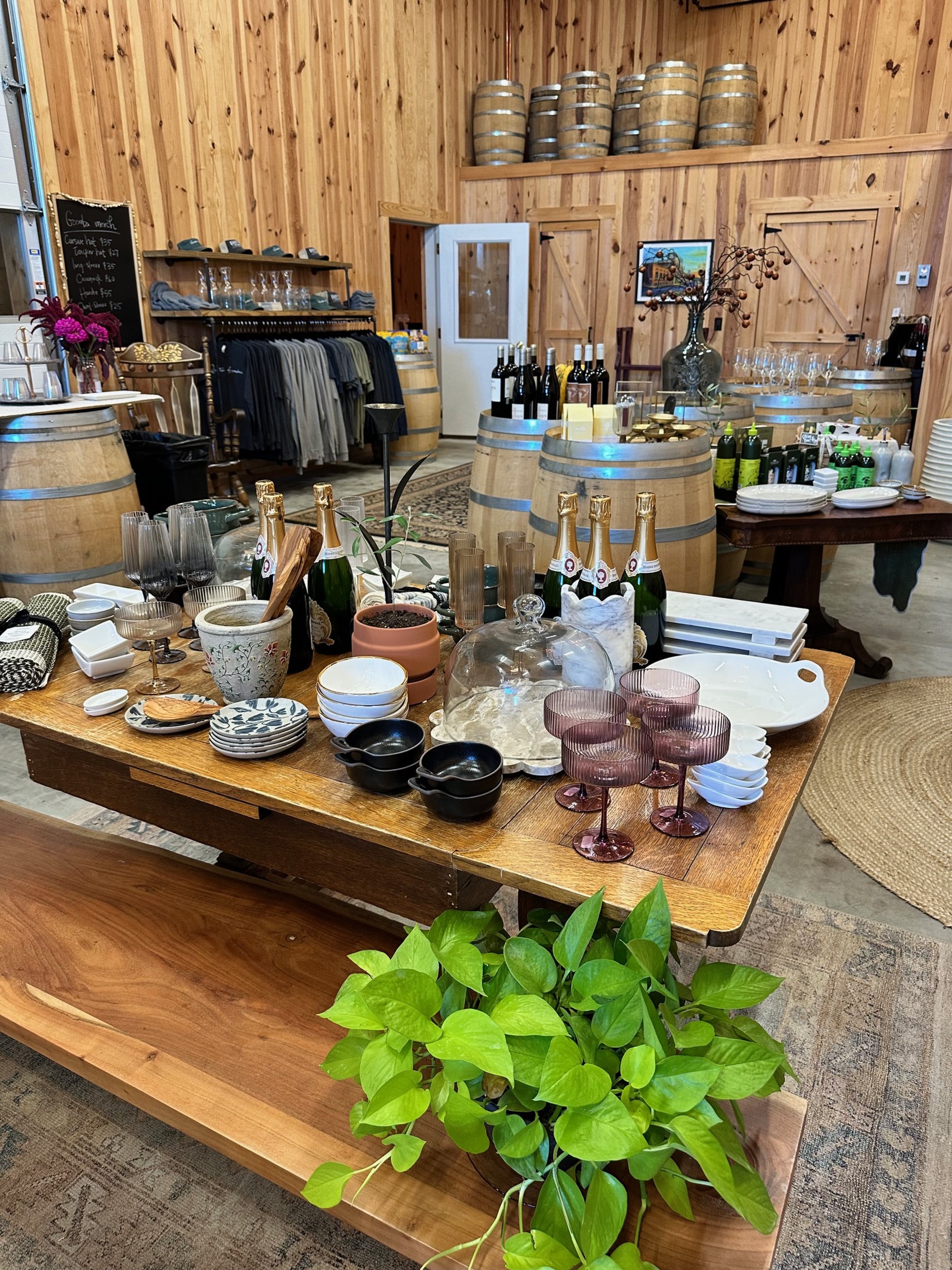 Interior of a rustic store or tasting room with wooden walls, displaying a variety of bottles, glassware, plates, and decor on tables and barrels, with a green potted plant in the foreground.