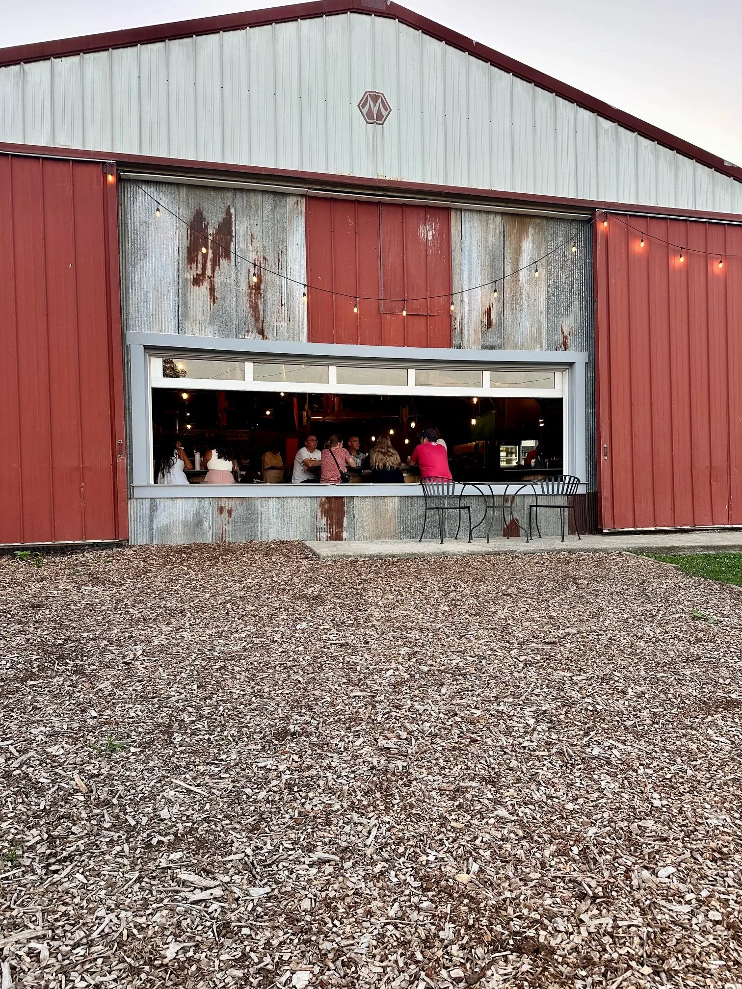 People sitting inside a restaurant seen through a large open window on a rustic barn exterior with string lights above, and wood chips on the ground outside.