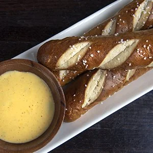 Two pretzel sticks with coarse salt served with a bowl of yellow mustard on a white rectangular plate.