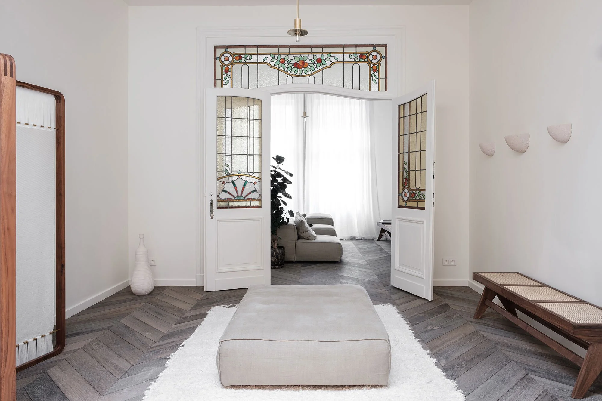 Woonkamer met glazen en houten dubbele deuren, een grijze poef op een wit vloerkleed, een houten bank, en een vaas in een hoek. De achterliggende ruimte heeft een grote kamerplant en een stoffen bank.