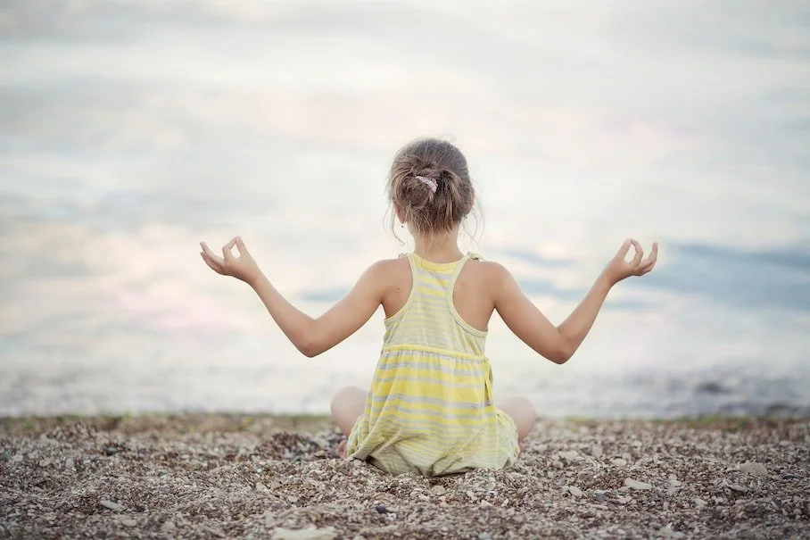 a human child in a yellow shirt sits in a meditative pose in the sunlight