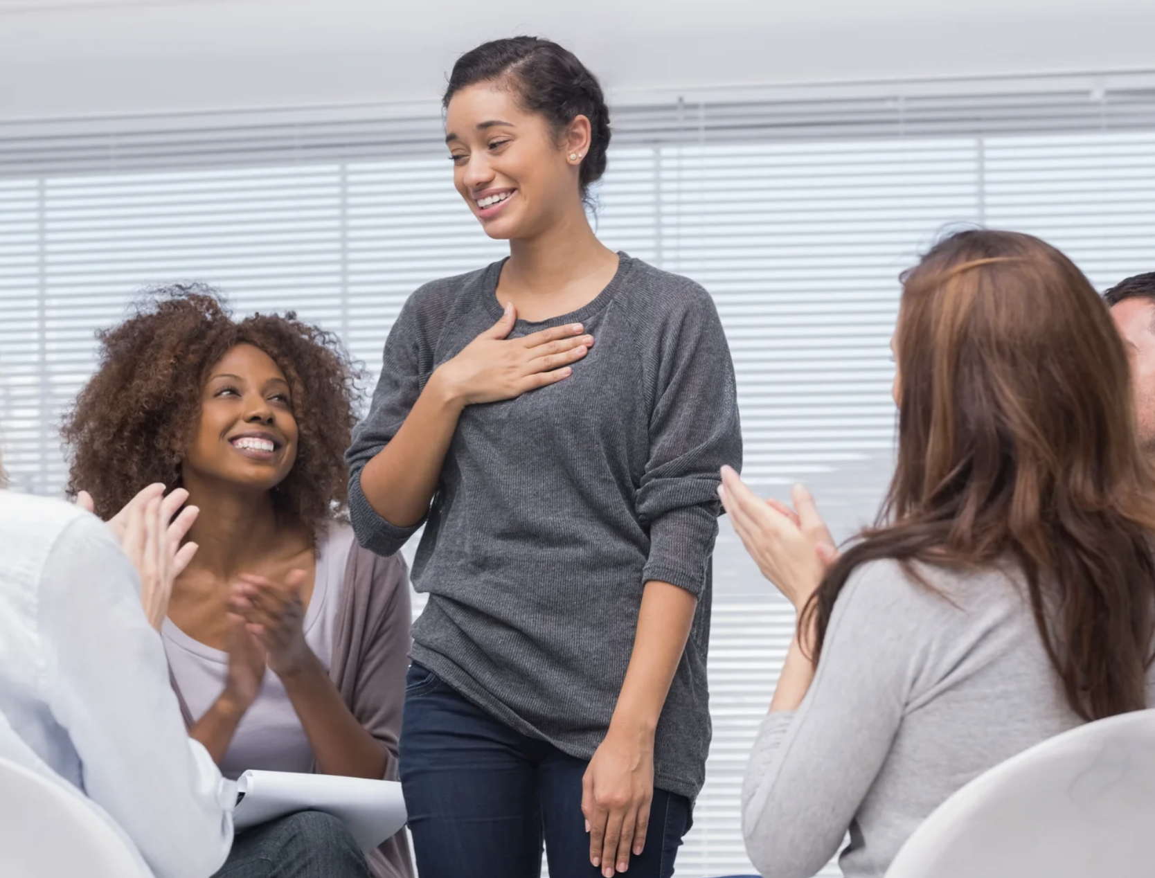 A human with light brown skin wearing a grey shirt puts their hand on their chest, as if relieved, as other humans clap for them