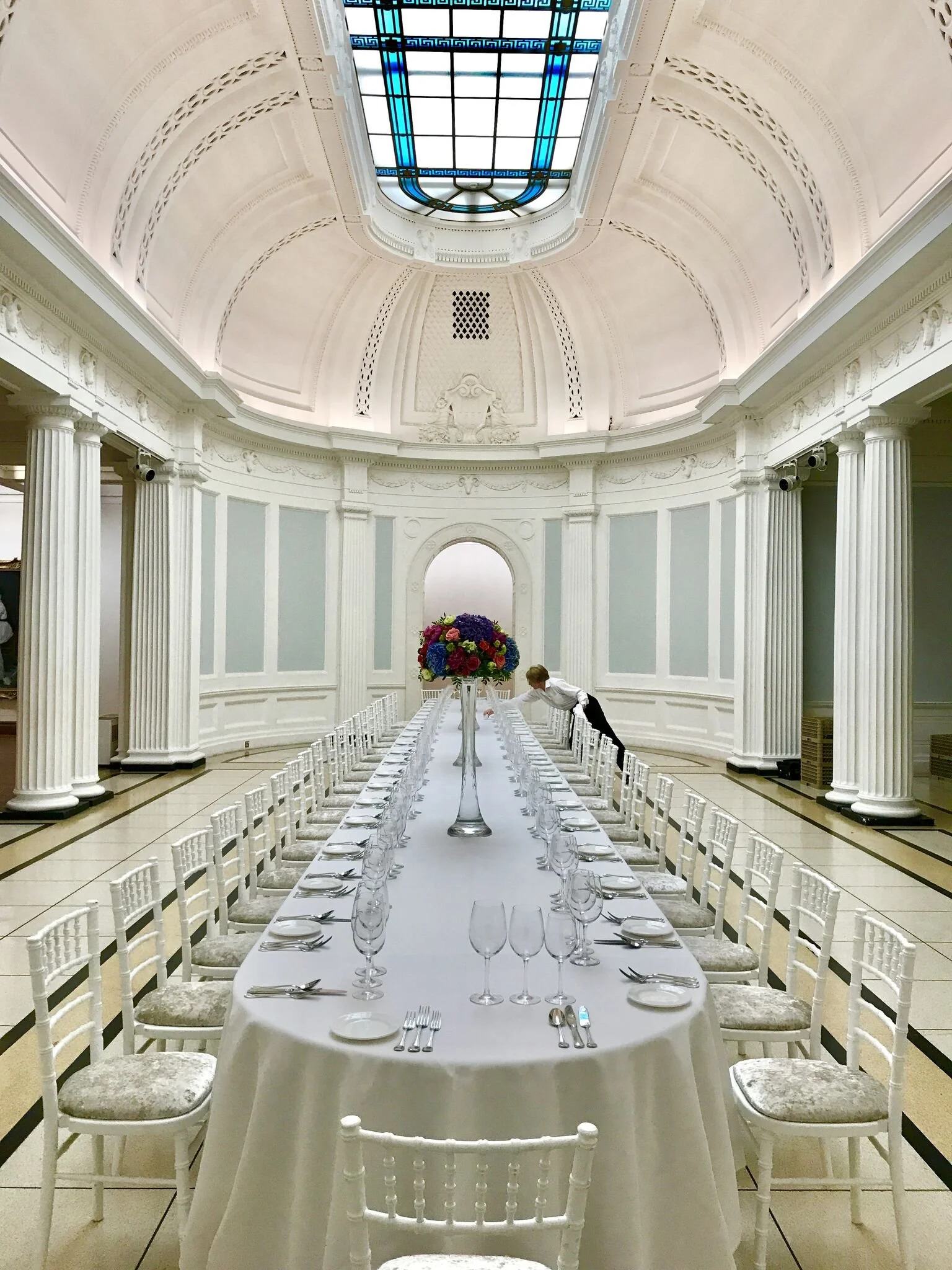 A long, elegantly set banquet table with white tablecloth, glasses, and silverware in a bright, grand room with a high ceiling, stained glass skylight, and white classical columns.