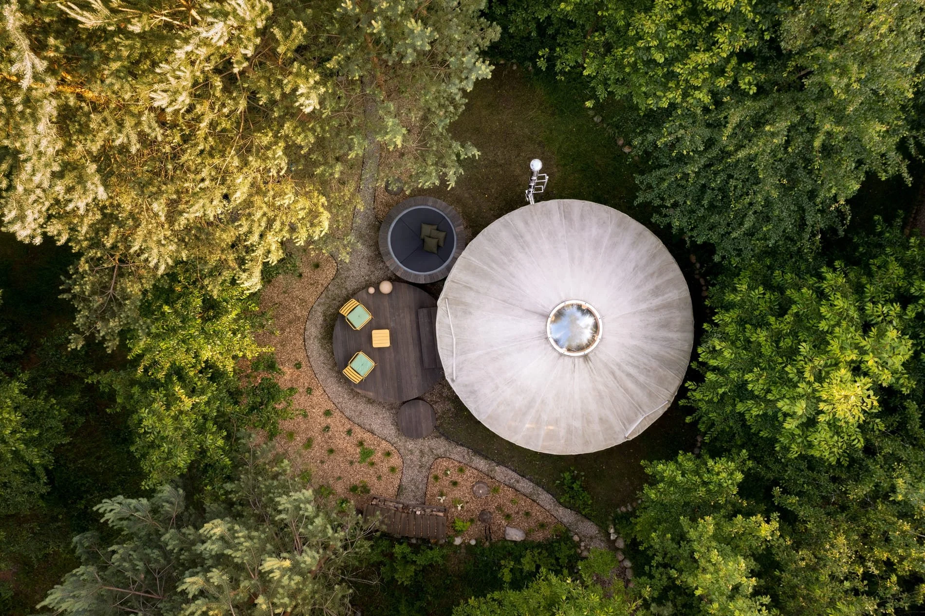 An aerial view of a backyard with a large circular patio, outdoor furniture including chairs and a round daybed with cushions, a large umbrella, surrounding lush green trees, and a gravel pathway.