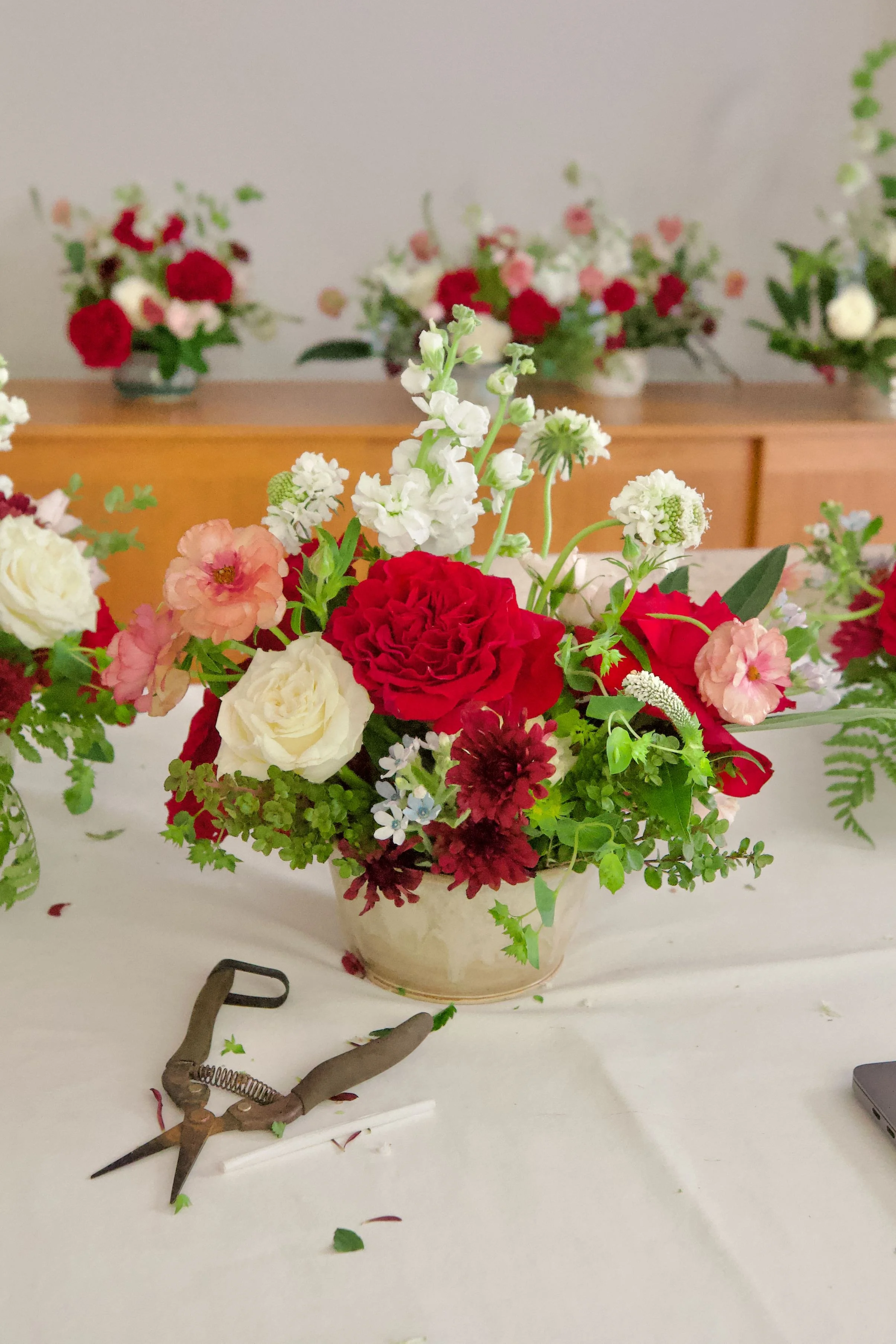 A floral arrangement in a beige bowl with red, white, pink, and dark red flowers, surrounded by green foliage, placed on a white table with flower arranging tools nearby. In the background, there are similar floral arrangements on a wooden surface.