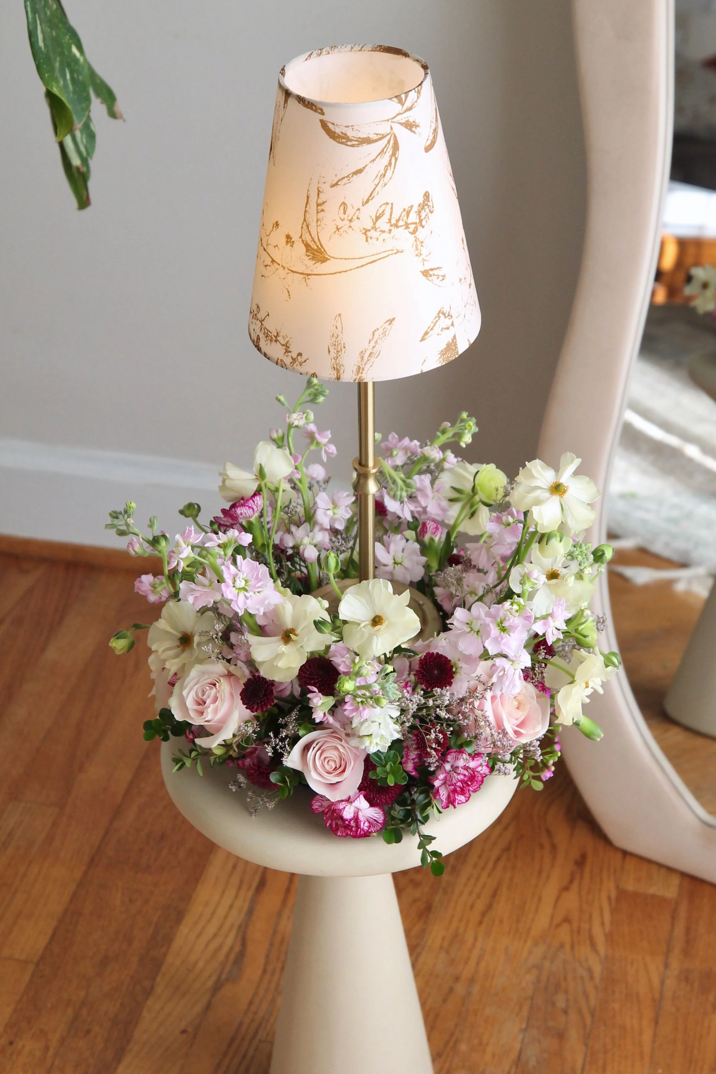 A table lamp with a floral-patterned shade, placed on a white pedestal table filled with pink and white flowers, including roses and other blossoms.