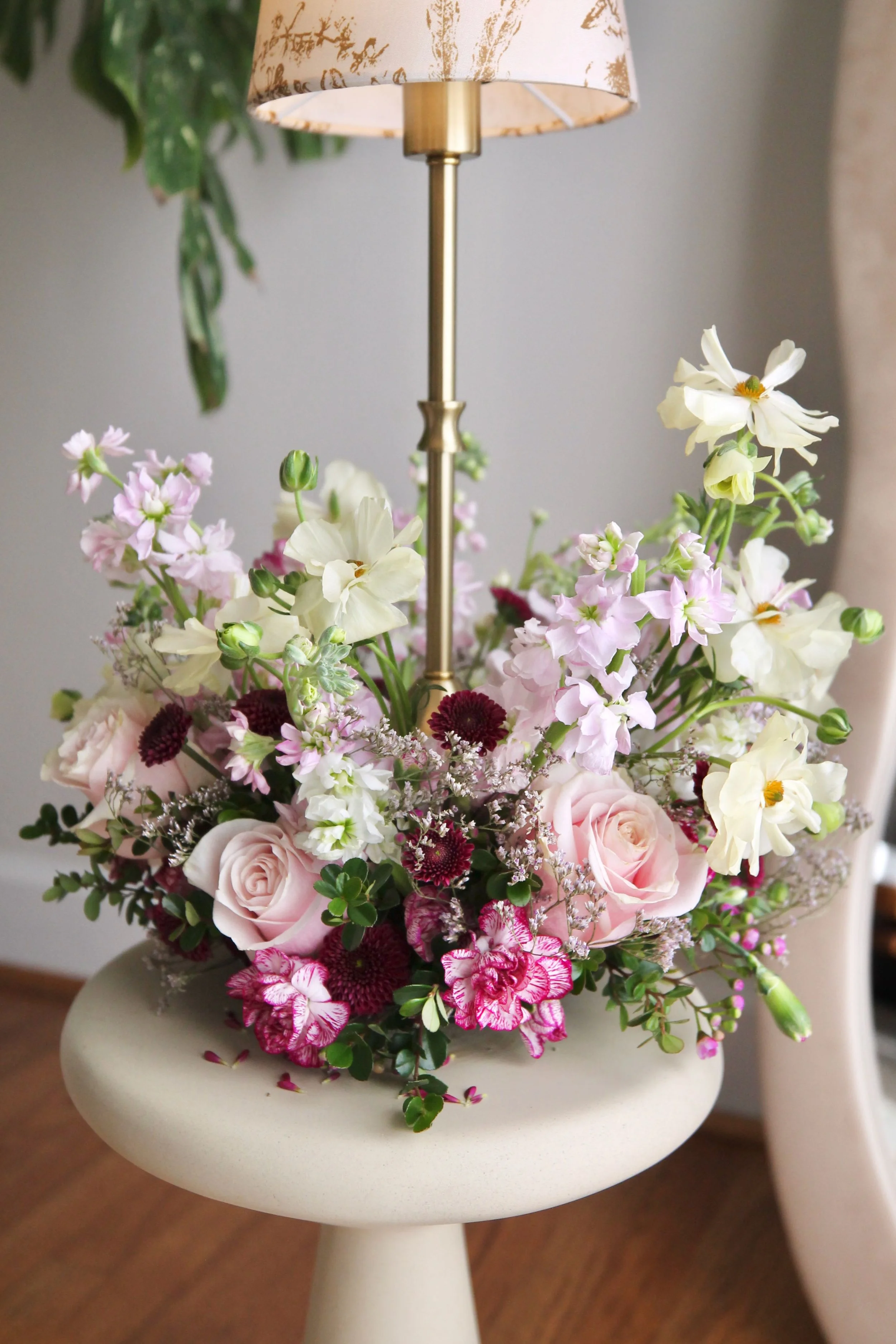 A floral arrangement with pink roses, white and pink carnations, and various other flowers, placed on a white pedestal stand with a lamp above it.