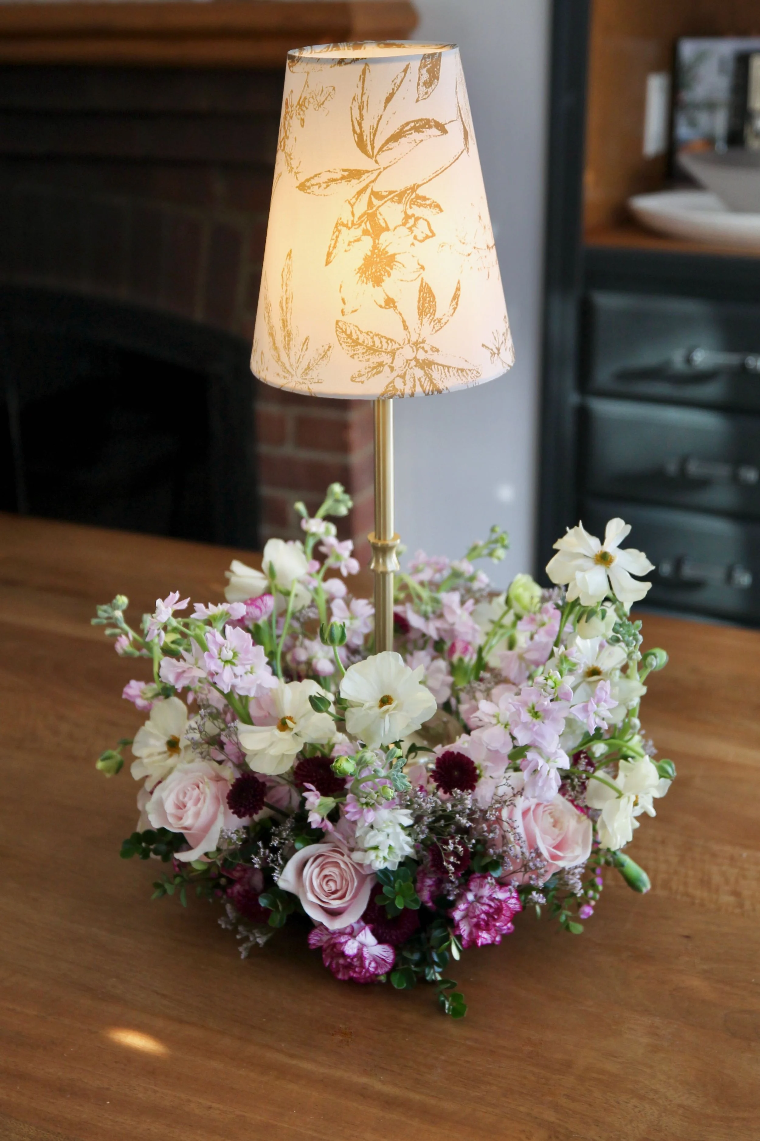 A flower arrangement with pink and white flowers placed on a wooden table, topped by a lamp with a floral-patterned lampshade.
