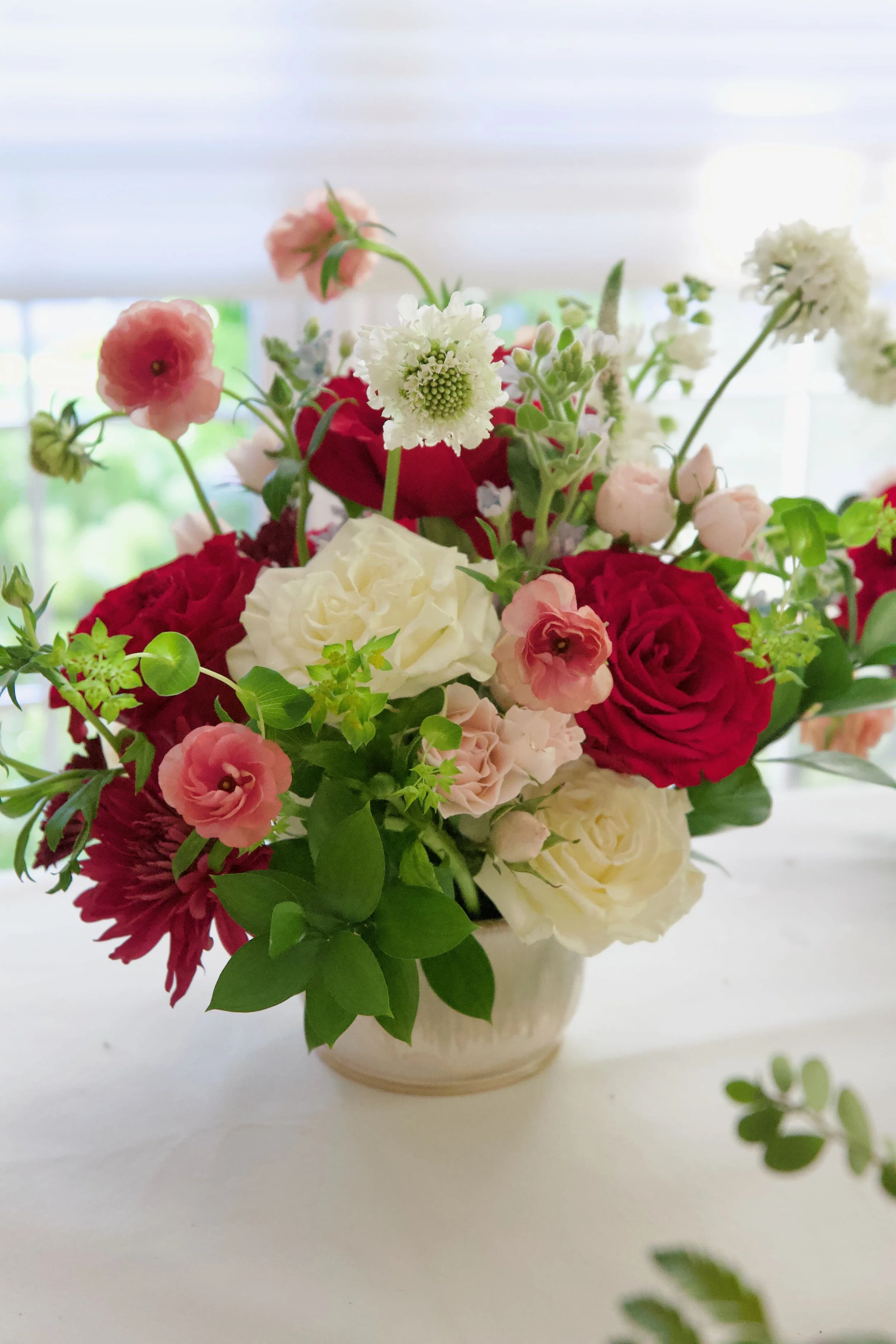 A floral arrangement with red, pink, white, and cream-colored flowers in a white ceramic vase on a white surface, with a blurred background of window blinds.