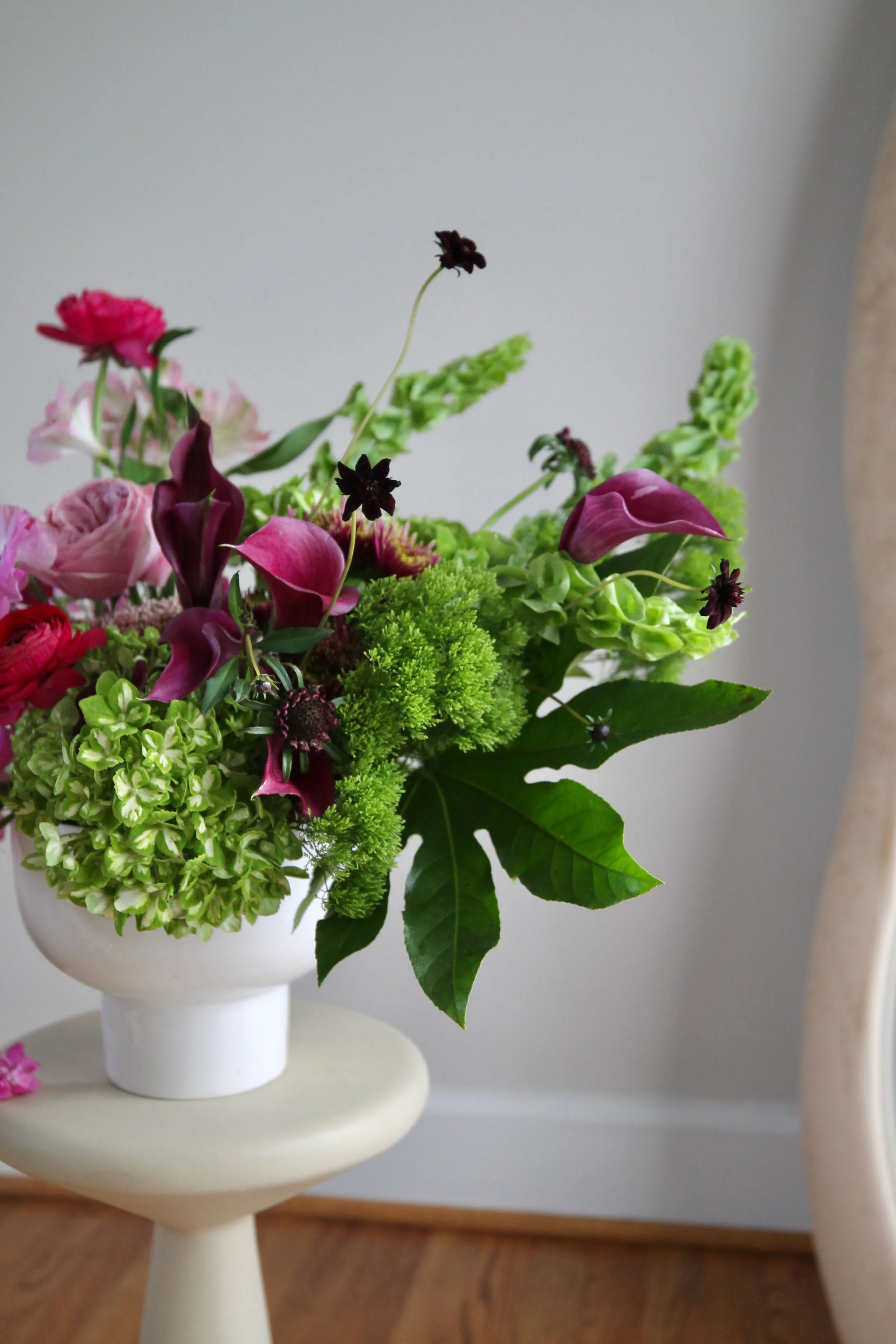 A floral arrangement with pink, purple, and green flowers in a white vase on a cream-colored stool.