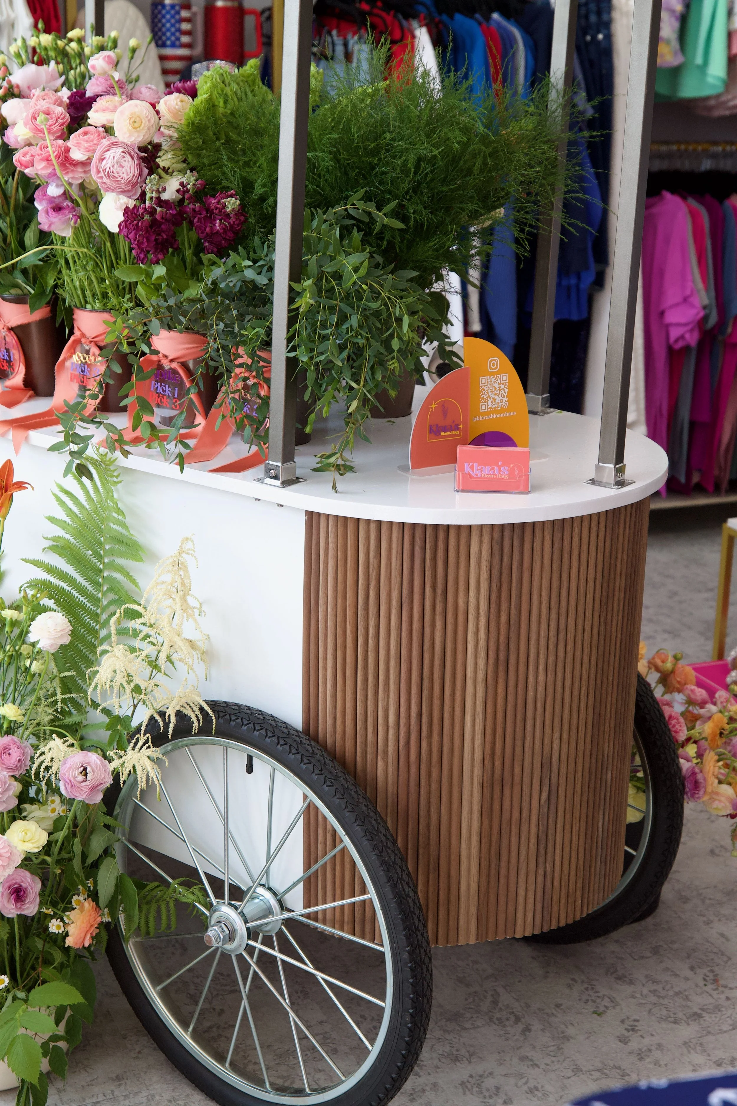 A flower cart with pink and purple flowers, green foliage, and a wooden lower section, located inside a store with clothing in the background.