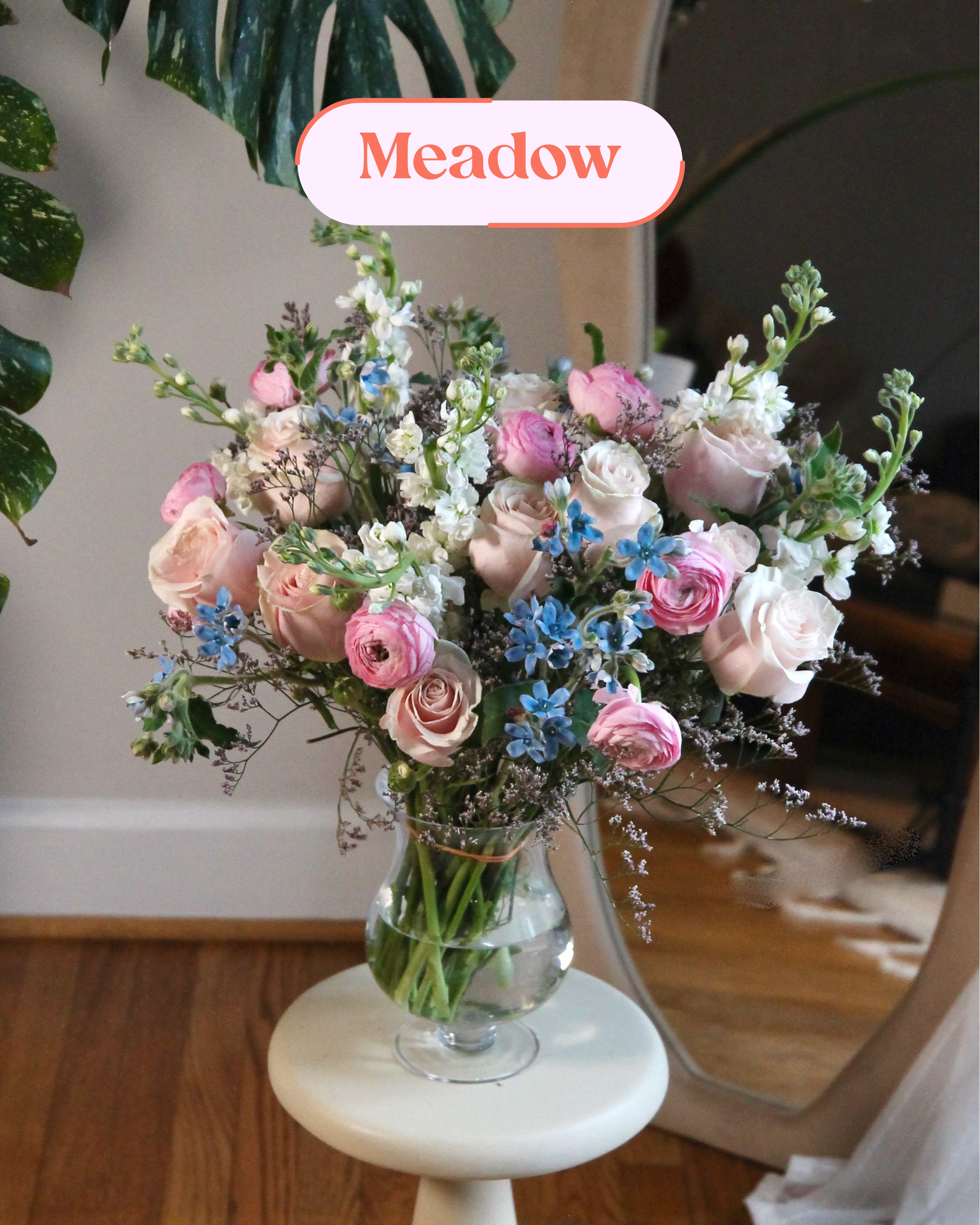 A large bouquet of pink, white, and blue flowers in a clear glass vase on a white stool.