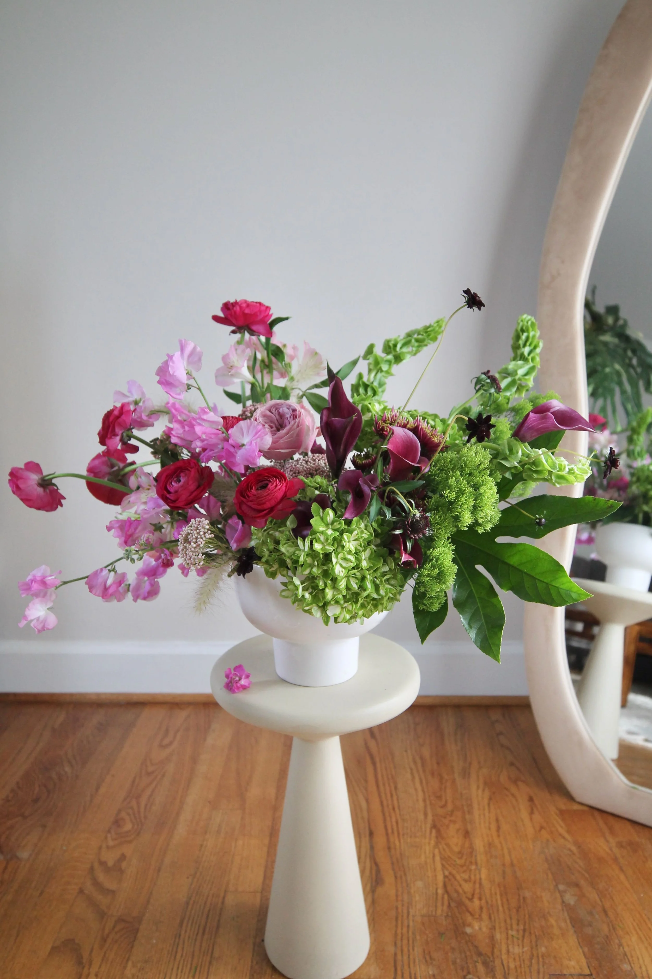 A floral arrangement with pink, purple, and green flowers in a white vase on a white pedestal table, reflected in a large mirror.