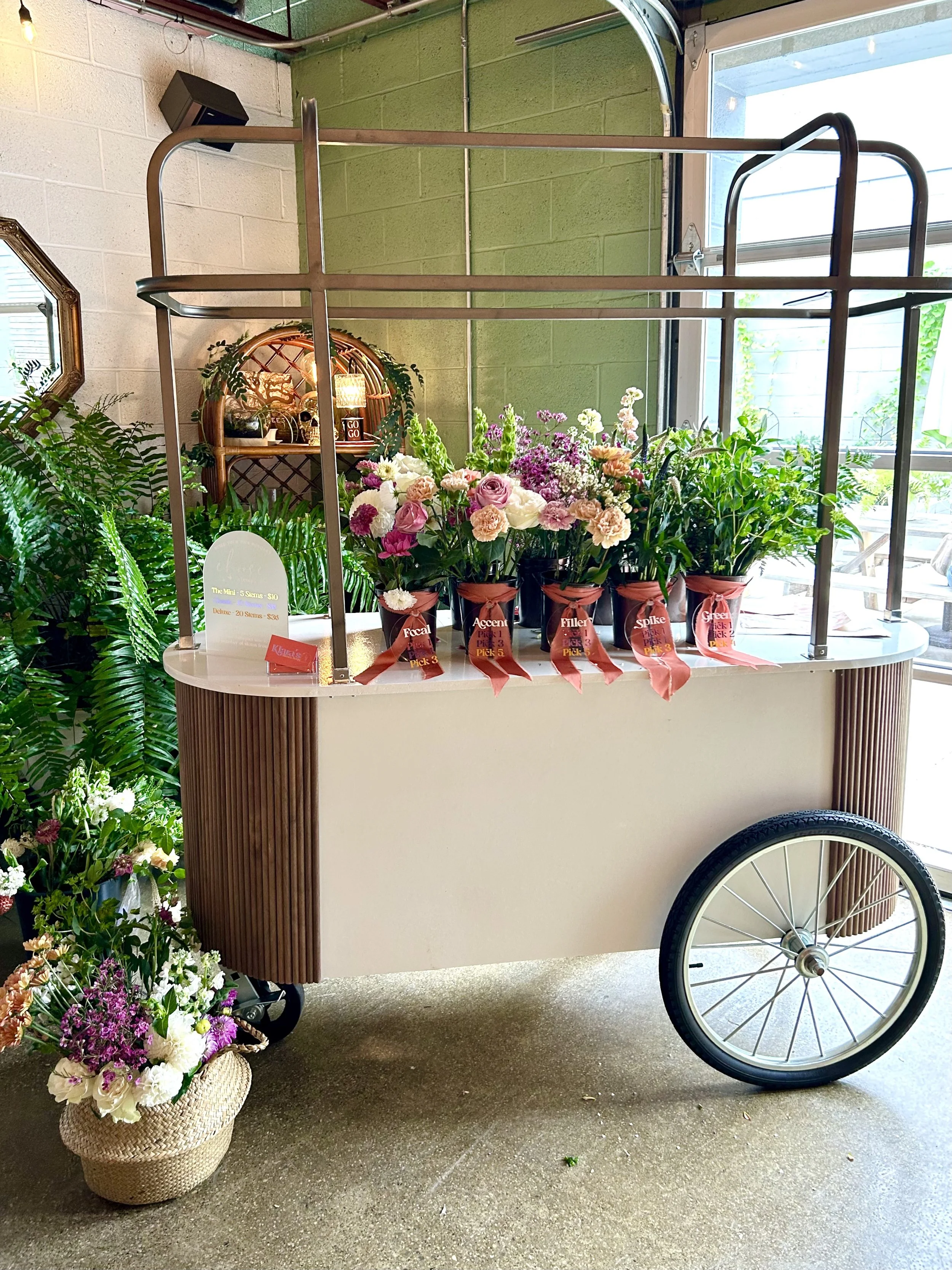 Flower cart with colorful flowers in pots on top and a basket of flowers on the floor beside it, located indoors near a large window at a private luxury event in Birmingham, MI.