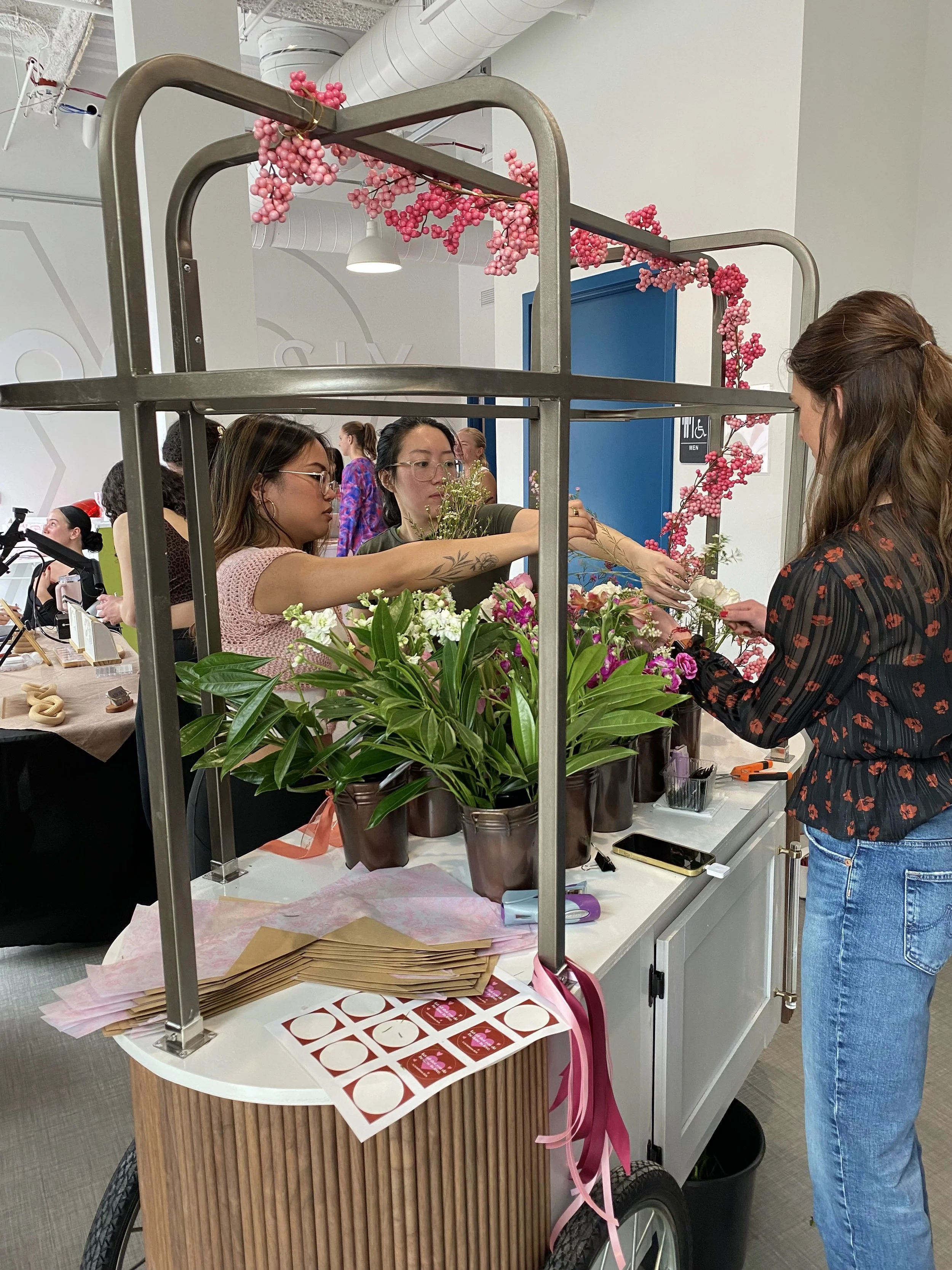 women at a floral shop, arranging pink and white flowers on a cart with potted plants and flower arrangements from a DIY Bouquet bar. A woman in a black shirt with red patterns and jeans is interacting with two women on the other side of the cart.