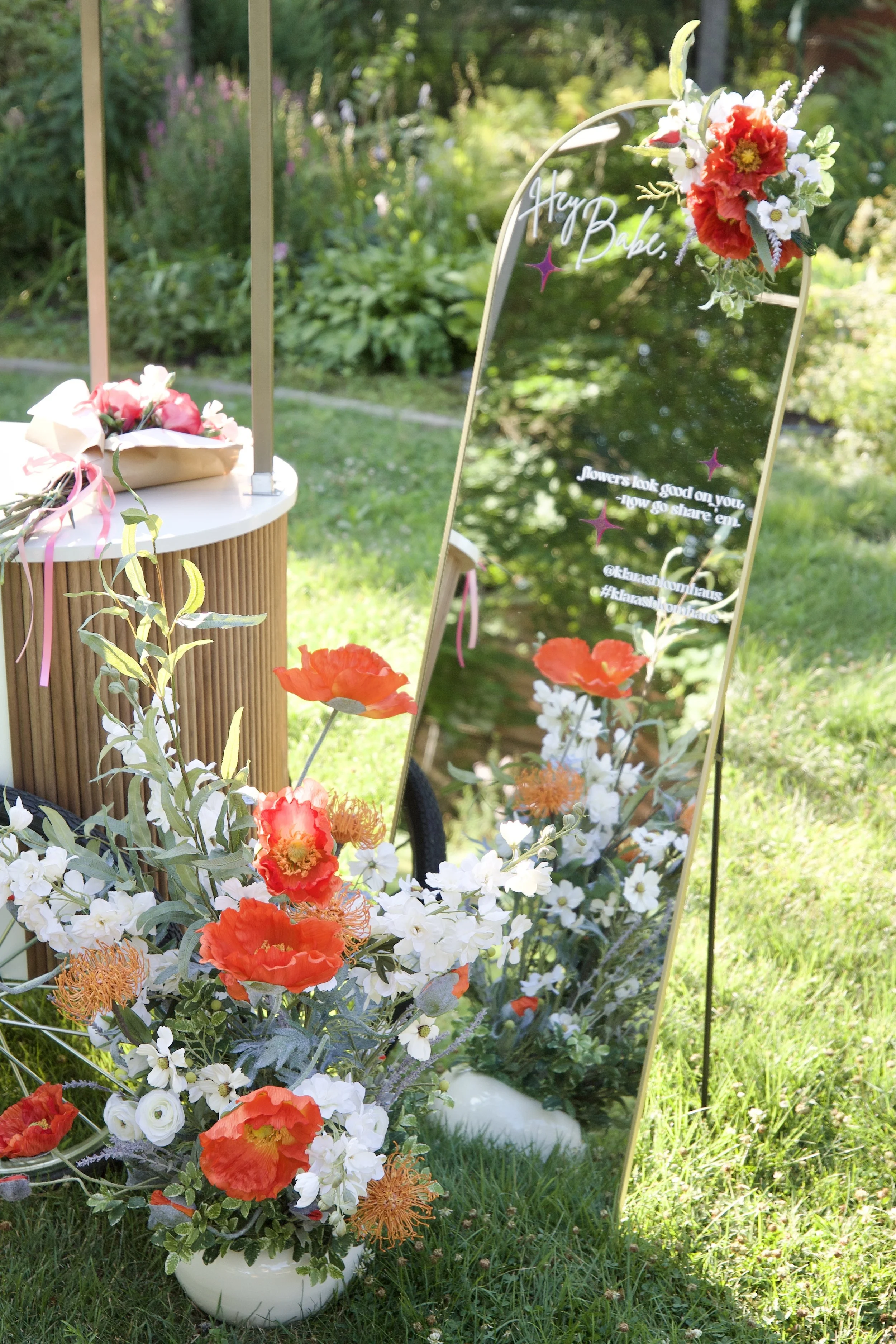 A tall, decorative mirror with a floral arrangement at the top corner, reflecting a garden scene, surrounded by a bouquet of orange and white flowers in a white vase, with a small table holding a wrapped gift nearby.