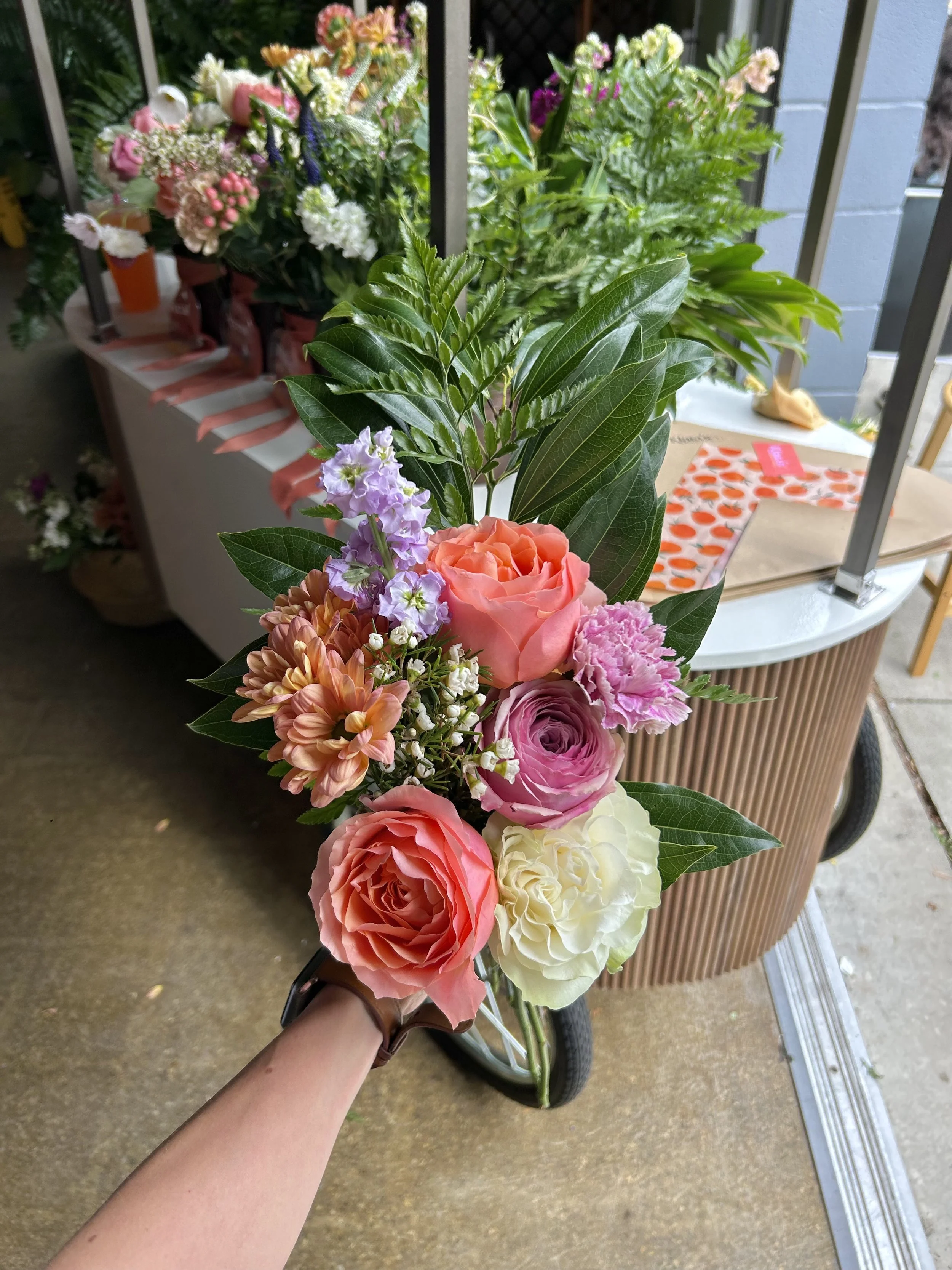Hand holding a bouquet of colorful flowers, including roses, carnations, and other blooms, with green leaves, in front of the Klara's Bloom Haus Metro Detroit flower cart in Royal Oak, MI.